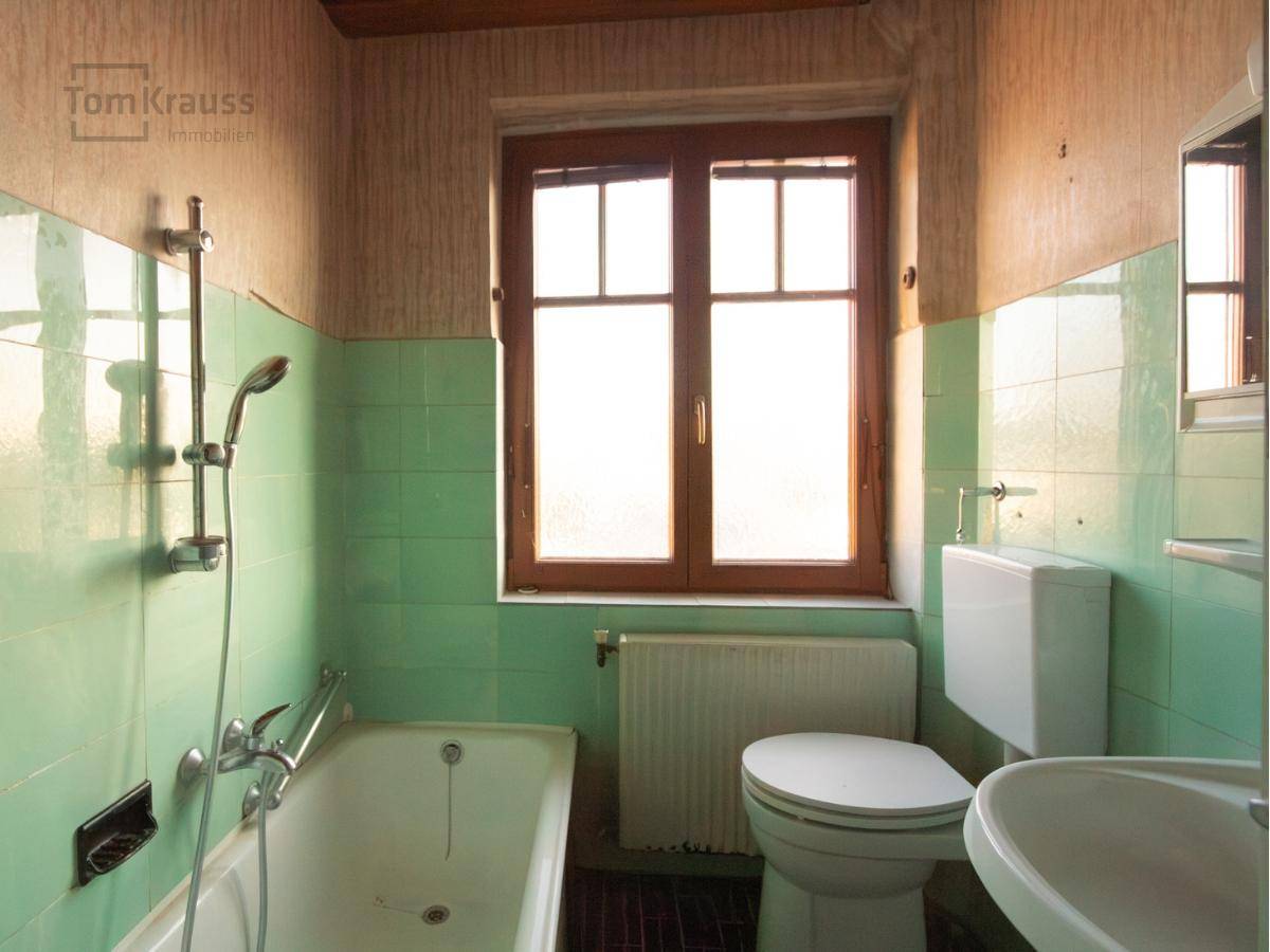 Badezimmer im Stil der 80er Jahre mit grünen Fliesen, Badewanne, Toilette und Holzfenster.