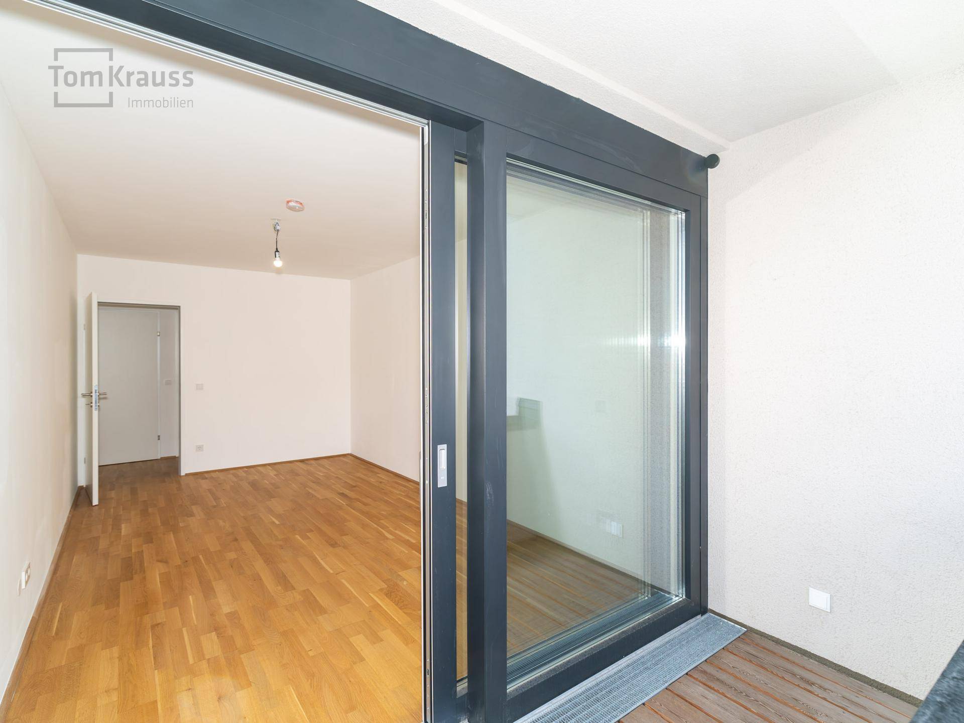 Heller, leerer Raum mit Parkettboden, gesehen durch die Schiebetüren eines Balkons.