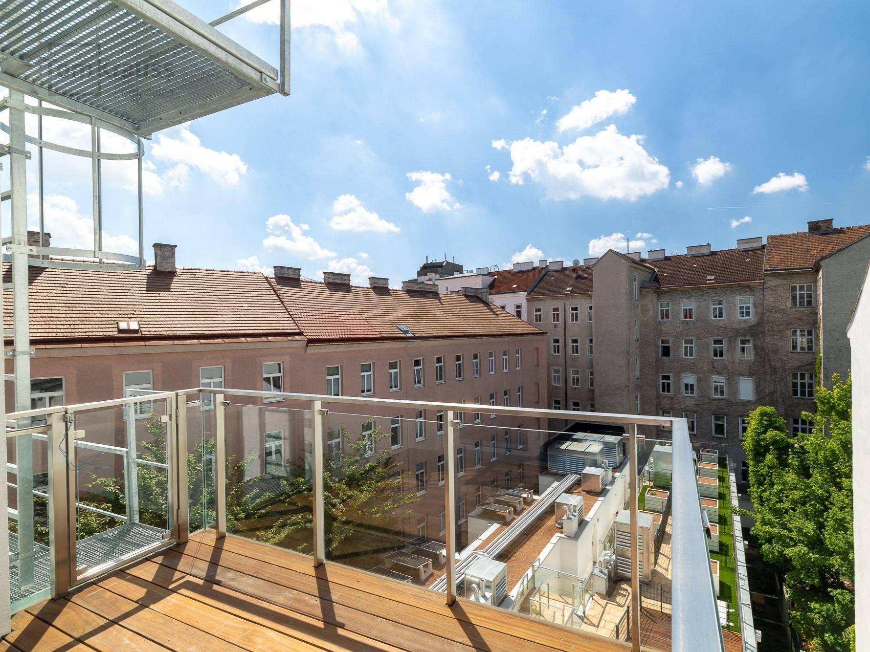 Moderner Balkon mit Holzbelag und Glasgeländer, bietet einen weiten Blick auf die umliegenden Gebäude.