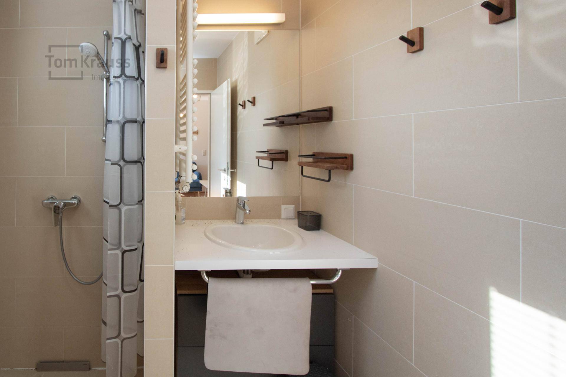 Badezimmer mit Dusche und Waschbecken, hell gefliest und mit praktischen Ablagen.