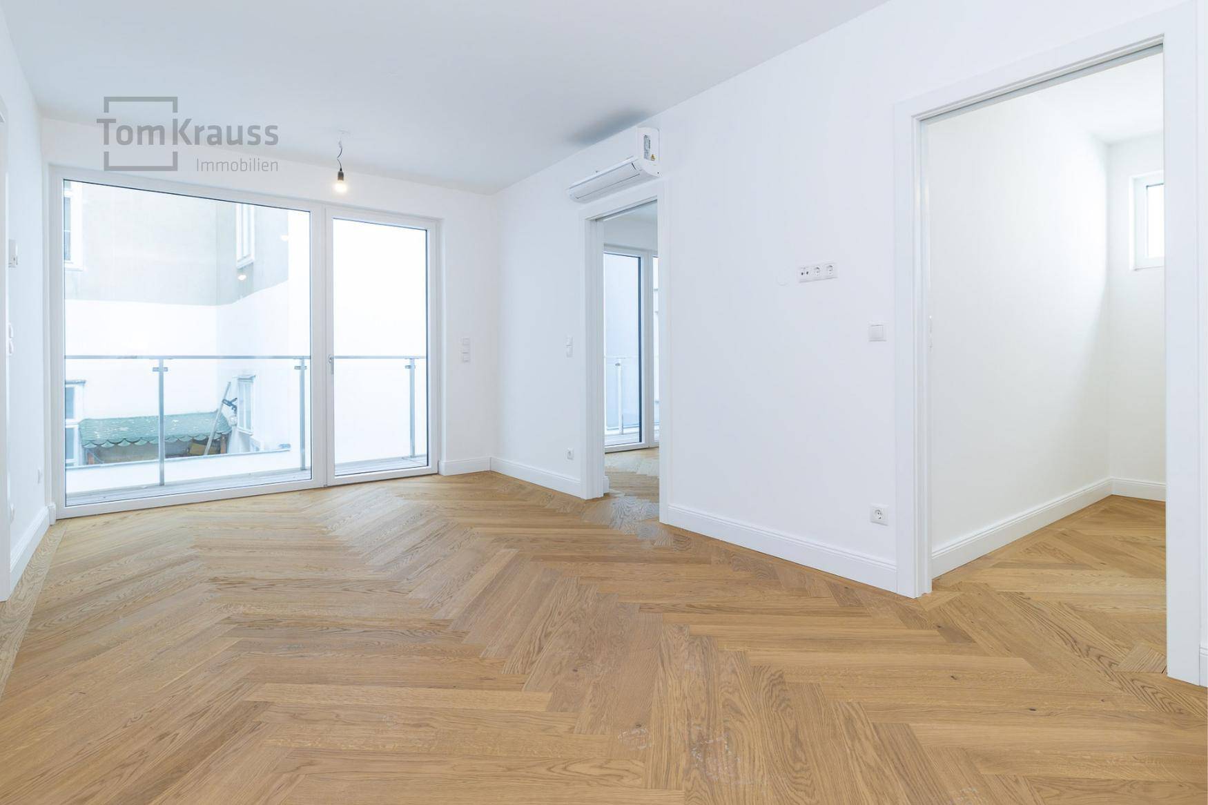 Helles, leeres Zimmer mit Fischgrätparkett, weißen Wänden und Zugang zu einem Balkon.
