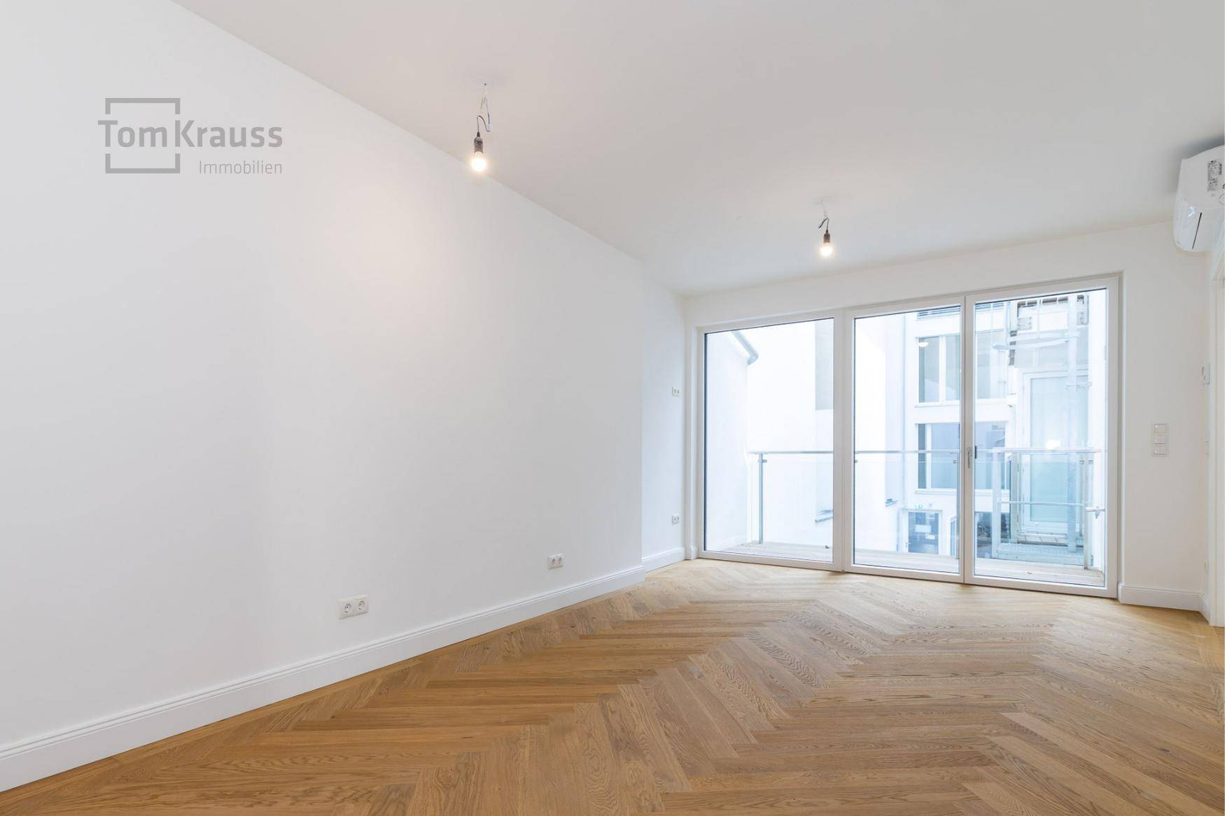 Geräumiges, unmöbliertes Zimmer mit hellem Fischgrätparkett und großen Fenstern zum Balkon.