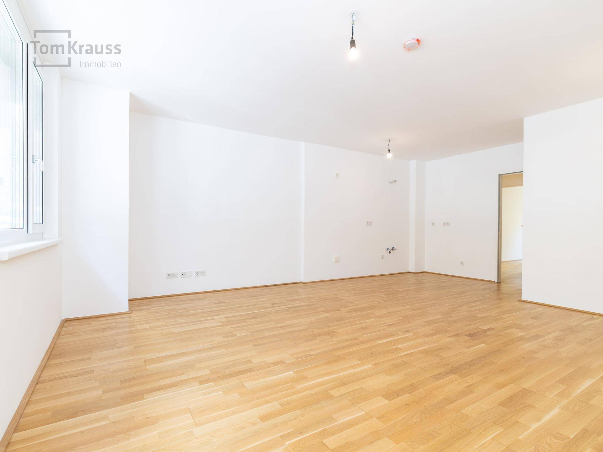 Leeres Zimmer mit Parkettboden und Fenstern, vorbereitet für die Installation einer Küche oder als Wohnbereich.