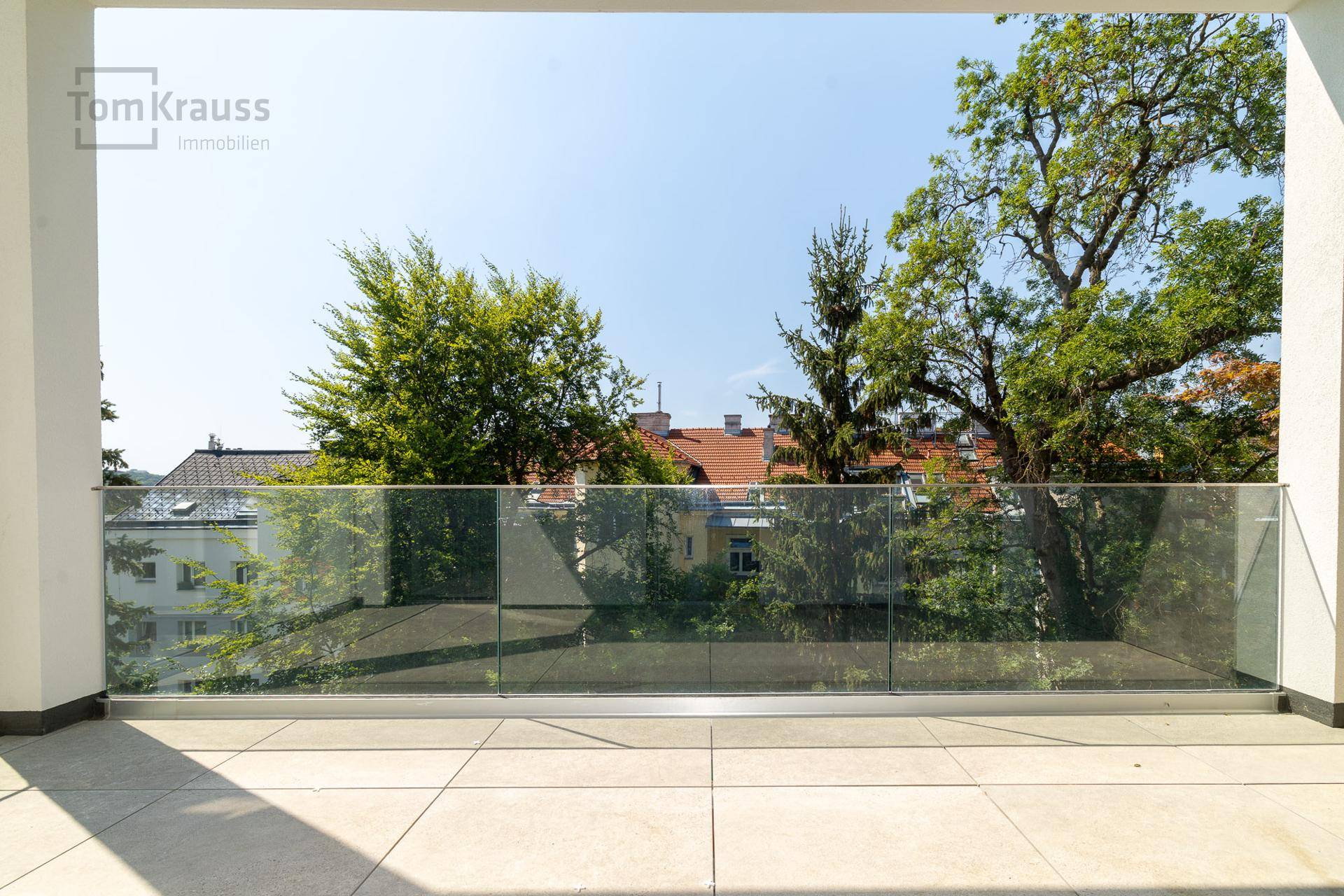 Geräumiger Balkon mit Fliesenboden und moderner Glasbrüstung, umgeben von Bäumen.