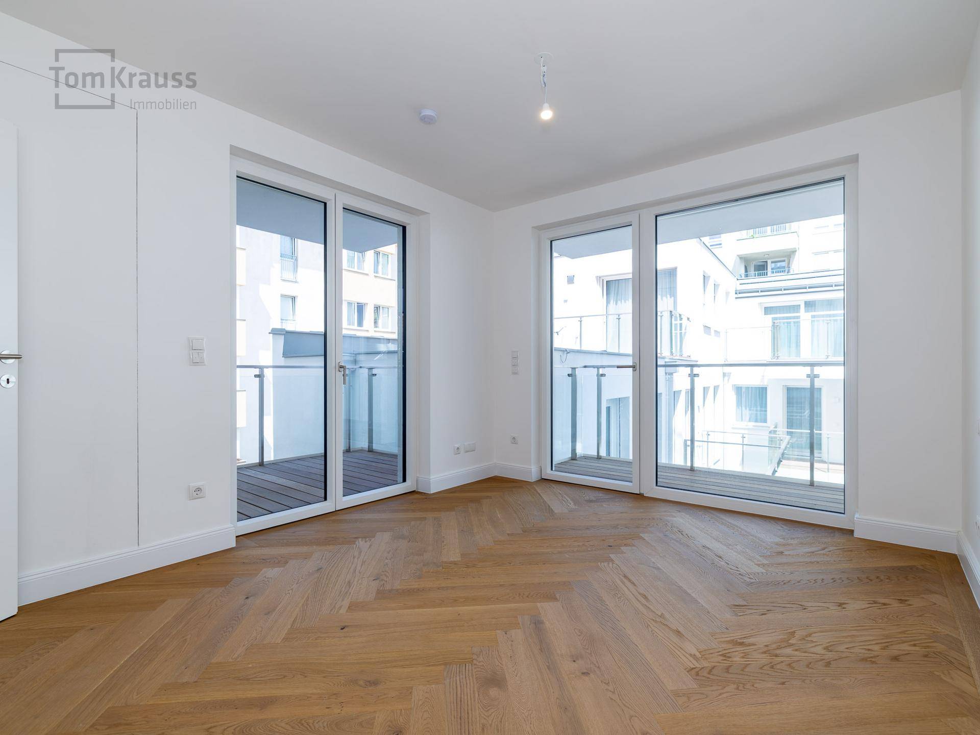 Helles, leeres Zimmer mit Fischgrätparkett und großen Fenstern, die auf einen Balkon führen.