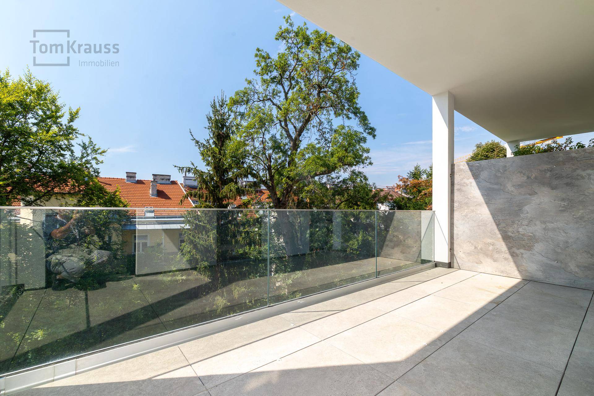 Geräumige Terrasse mit Fliesenboden und moderner Glasbrüstung, umgeben von Bäumen.
