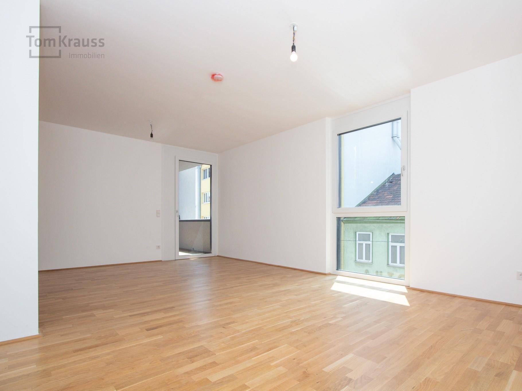 Heller, leerer Raum mit Parkettboden, großem Fenster und Zugang zu einem Außenbereich.