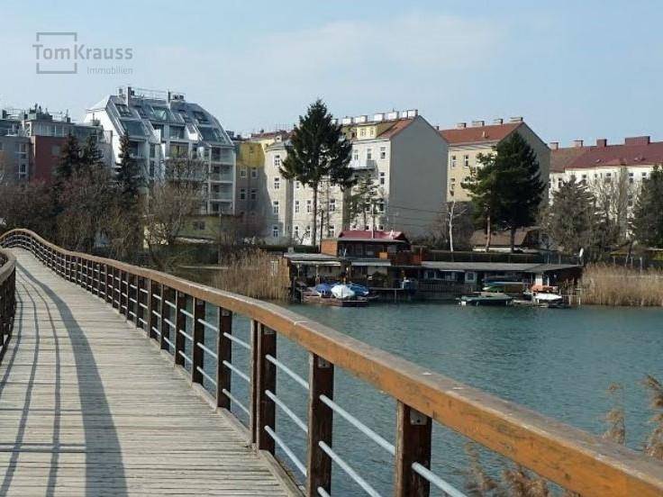 Holzbrücke über Wasser mit Gebäuden und Uferlandschaft im Hintergrund.