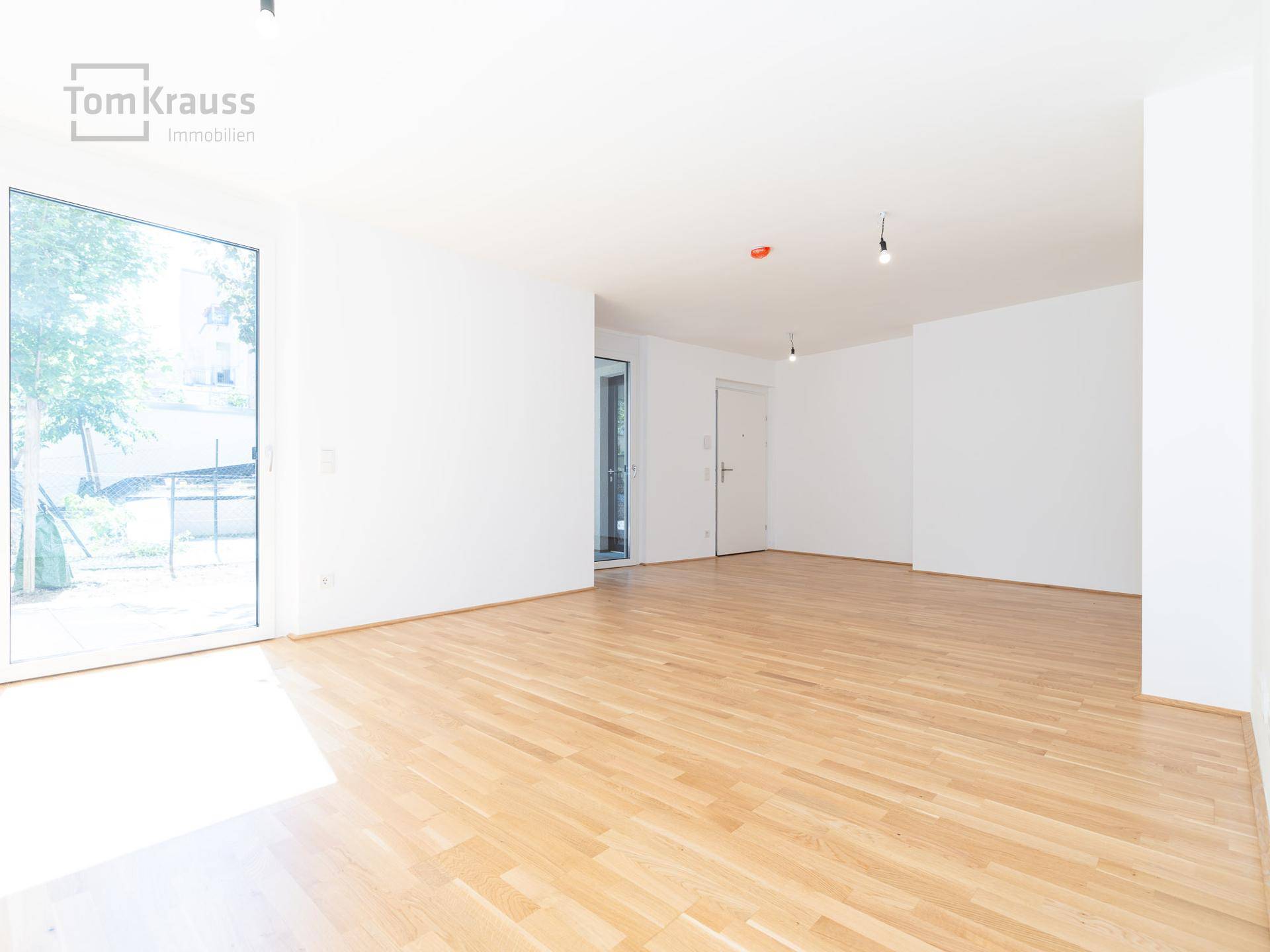 Heller, leerer Wohnbereich mit Holzboden und großer Fensterfront, die viel Tageslicht hereinlässt.