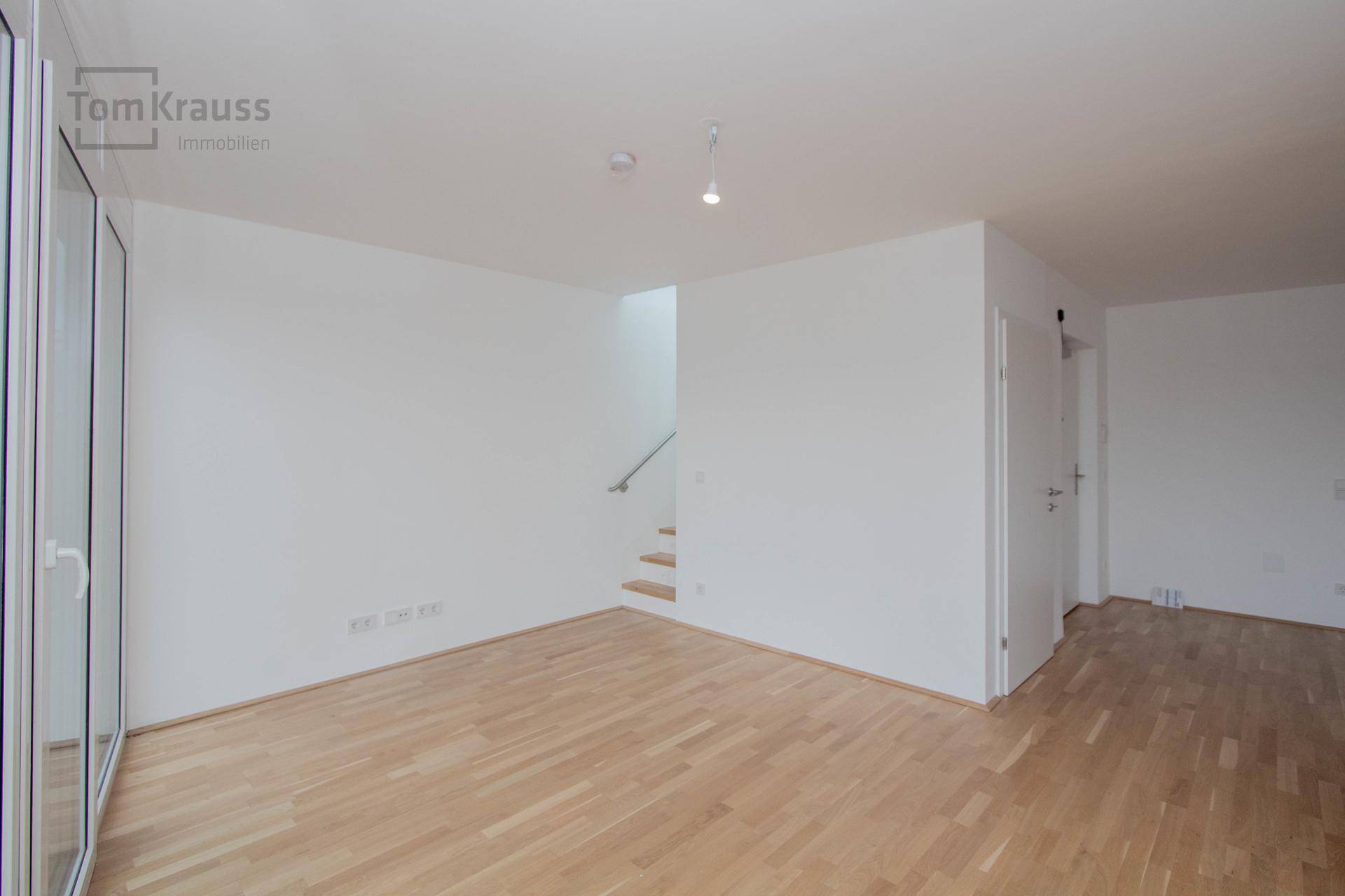 Heller Raum mit Holzboden, weißen Wänden und einer Treppe, die in die obere Etage führt.
