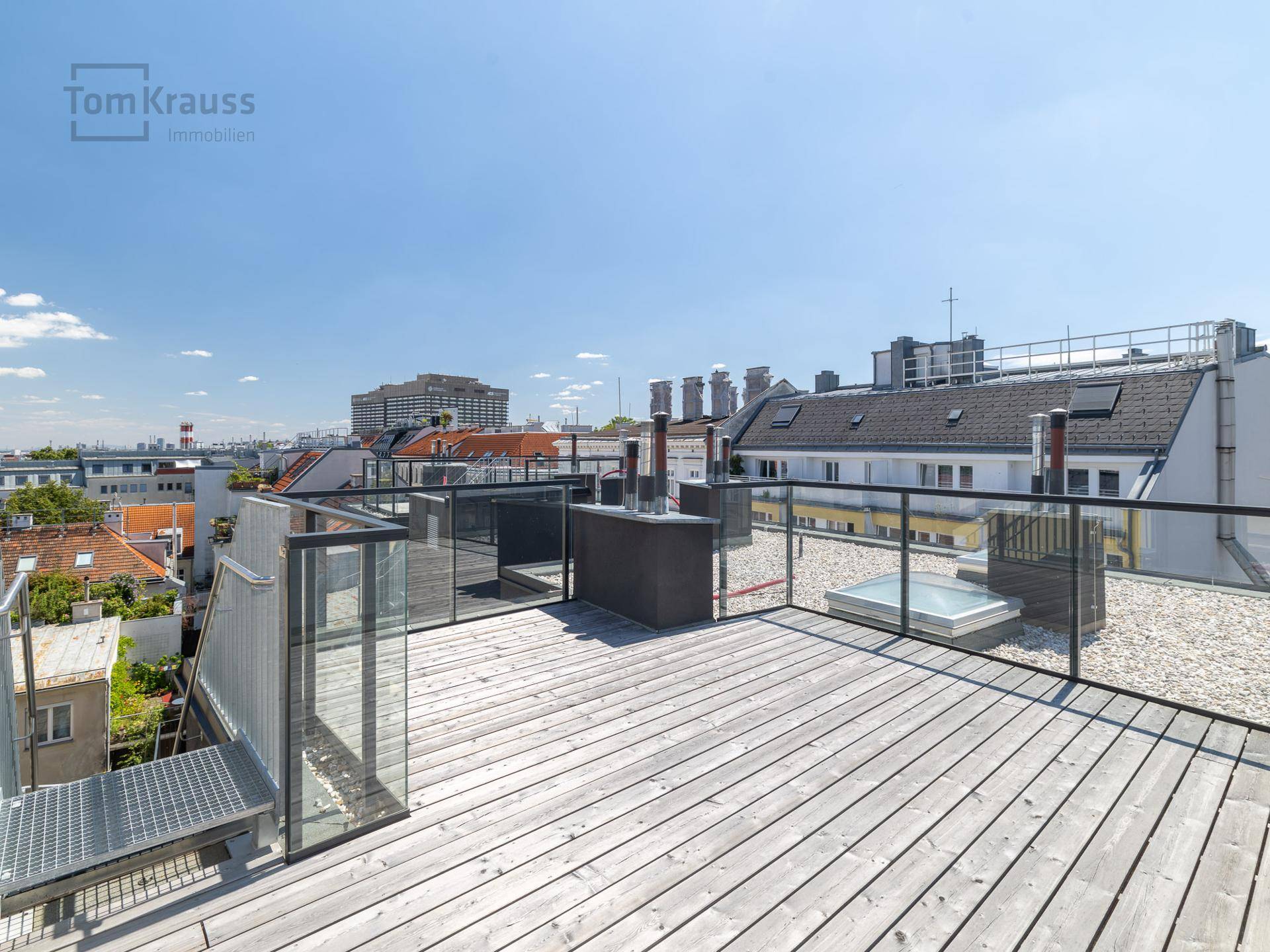 Geräumige Dachterrasse mit Holzboden, Glasgeländer und integriertem Whirlpool unter blauem Himmel.