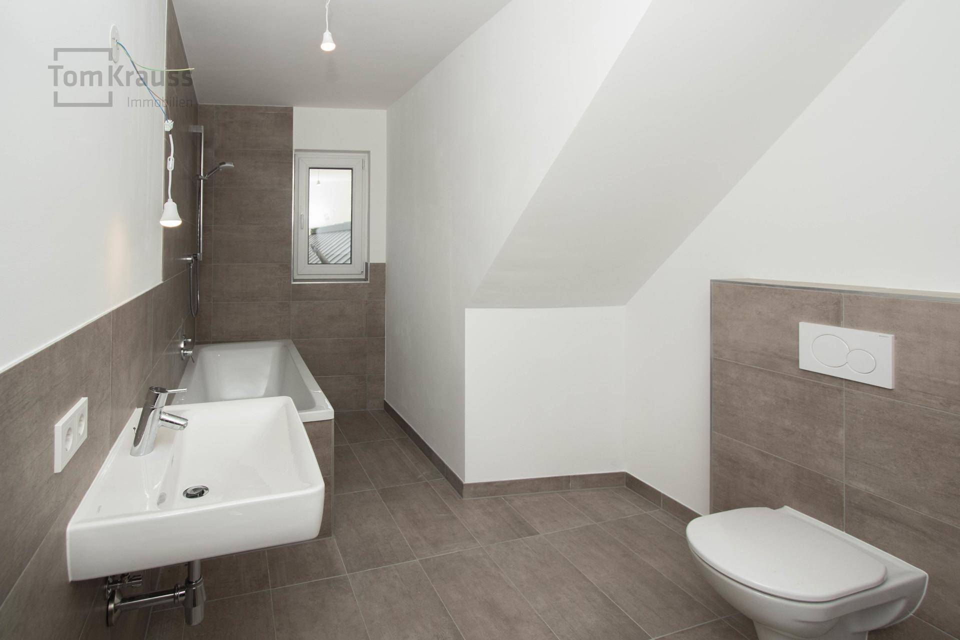 Helles Badezimmer mit Badewanne, Waschbecken, Toilette und grauen Fliesen unter einer Dachschräge.