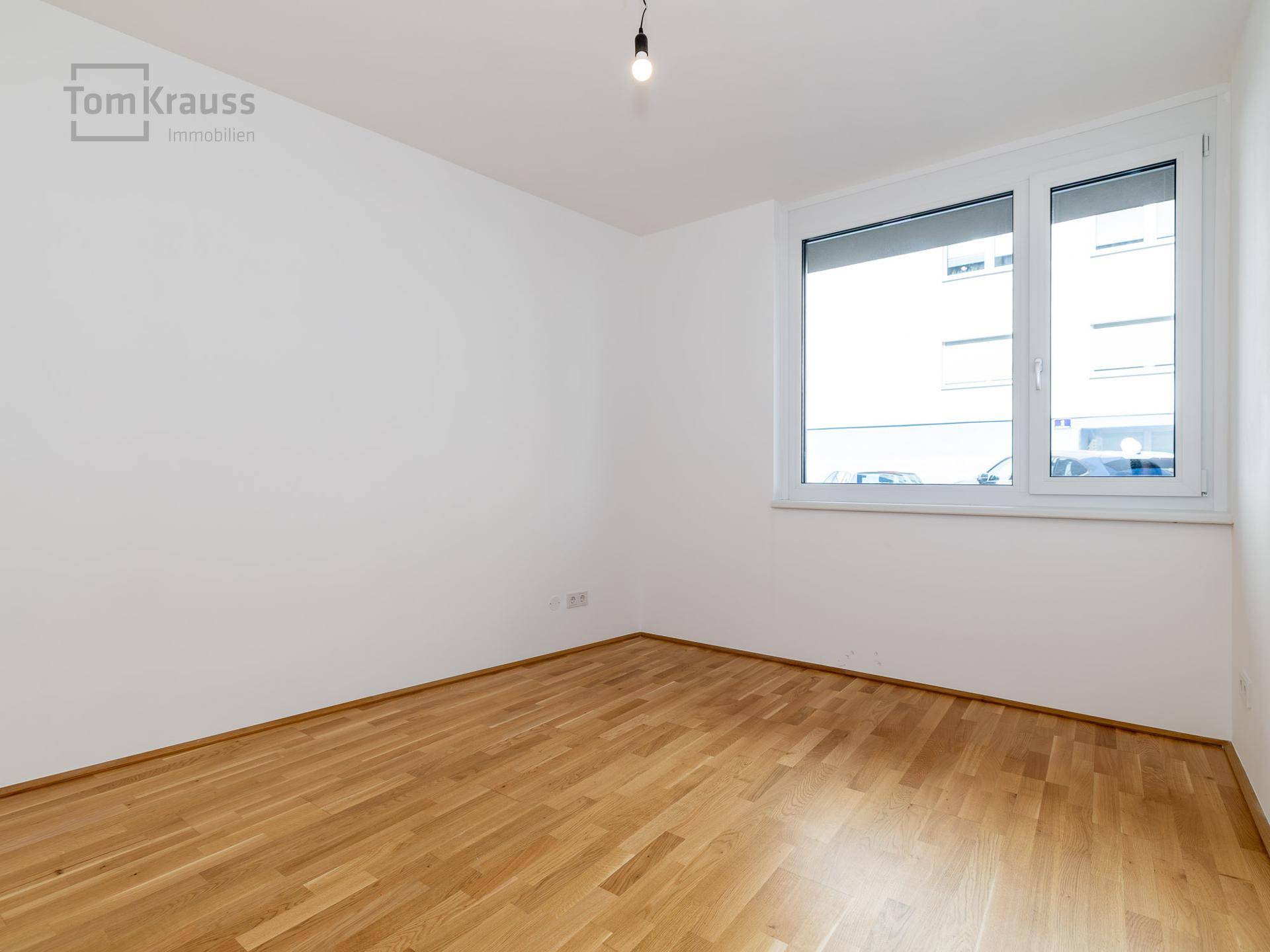 Leeres Zimmer mit hellem Holzboden und einem großen Fenster, das viel Tageslicht hereinlässt.
