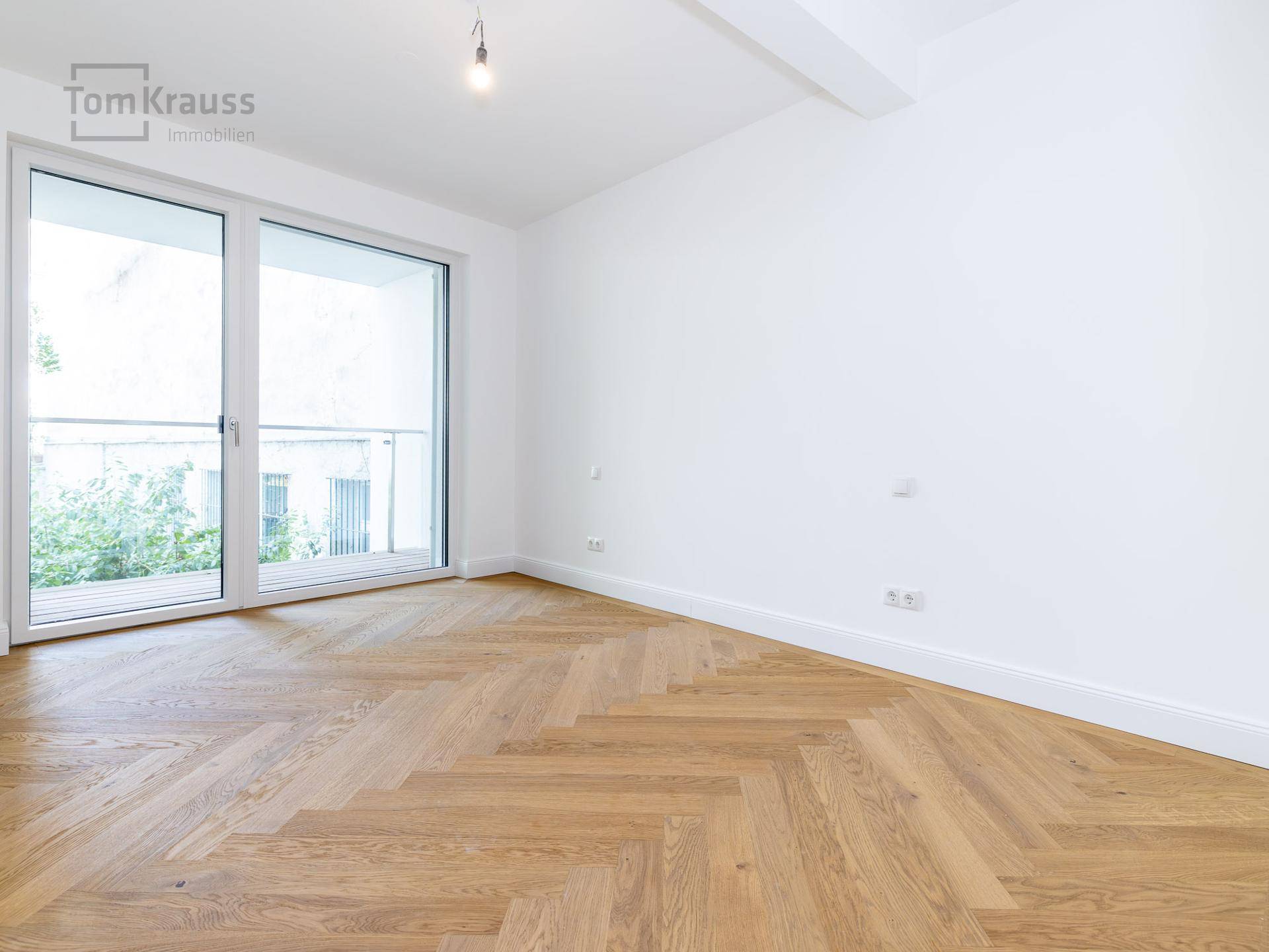 Leeres Schlafzimmer mit Fischgrätparkett und großen Fenstern zum Balkon.