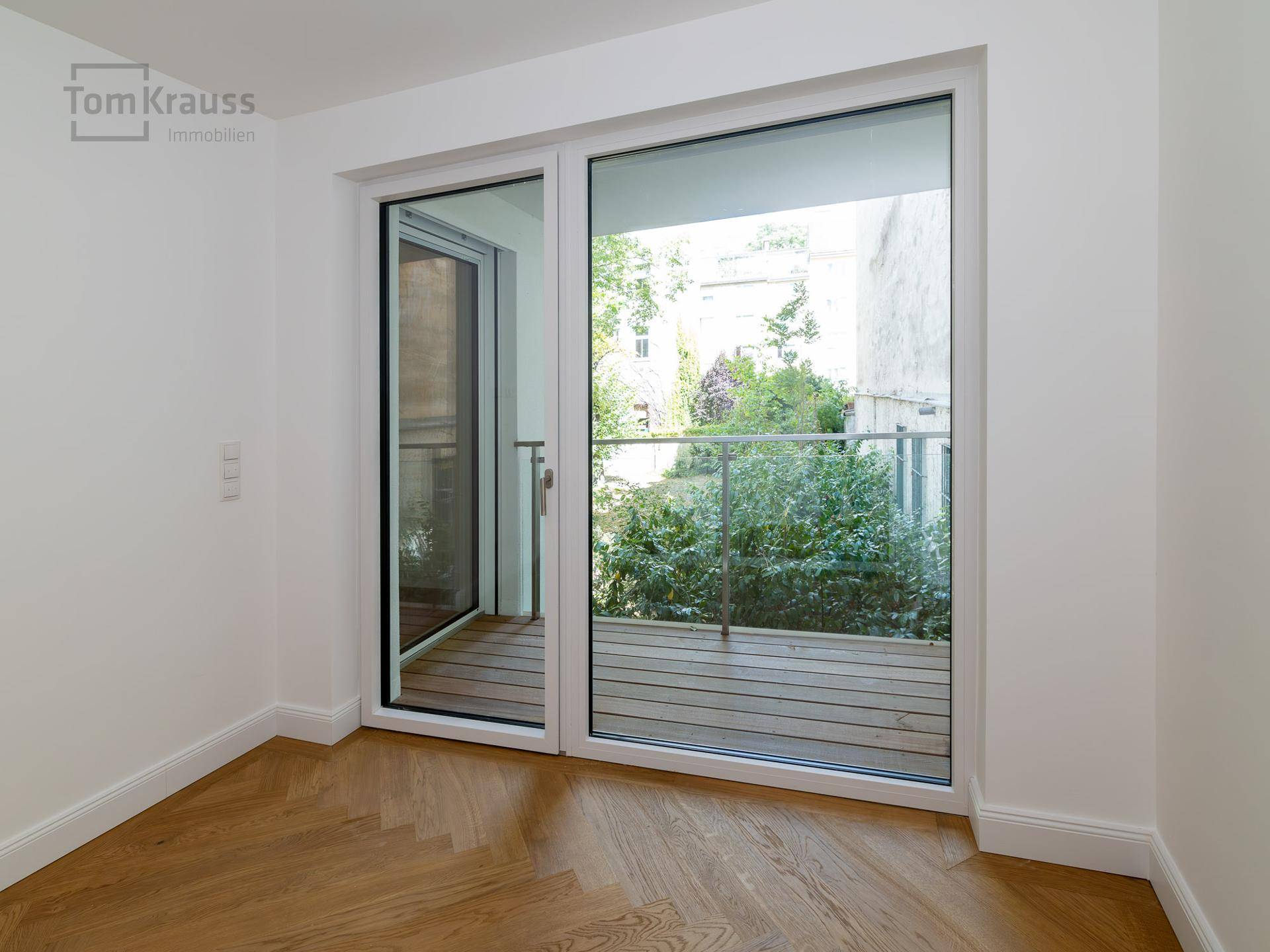 Heller Raum mit großen Fenstern und Zugang zu einem Balkon mit Holzbelag.