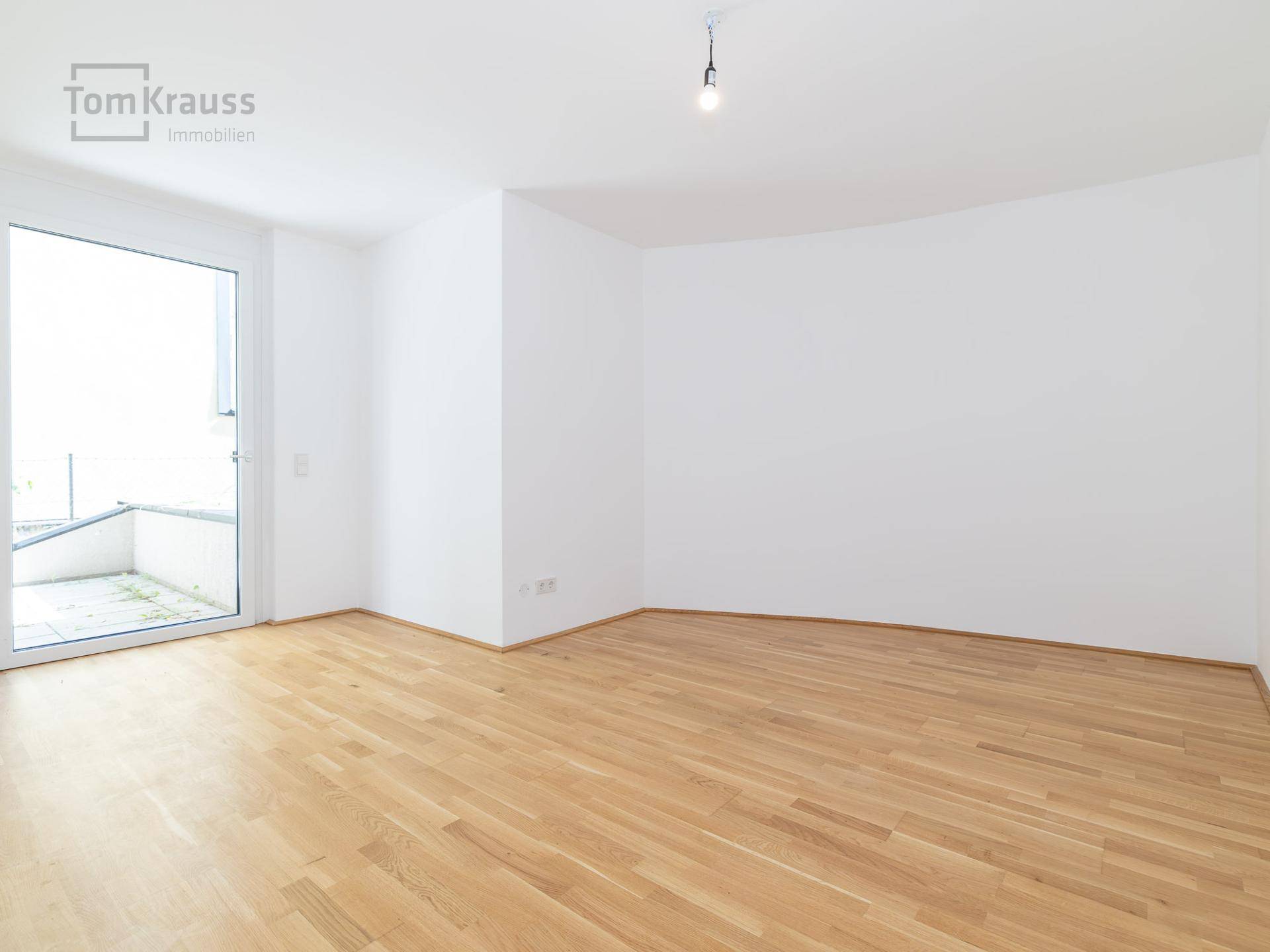 Leeres Zimmer mit hellem Parkettboden und großem Fenster mit Zugang zu einem Außenbereich.