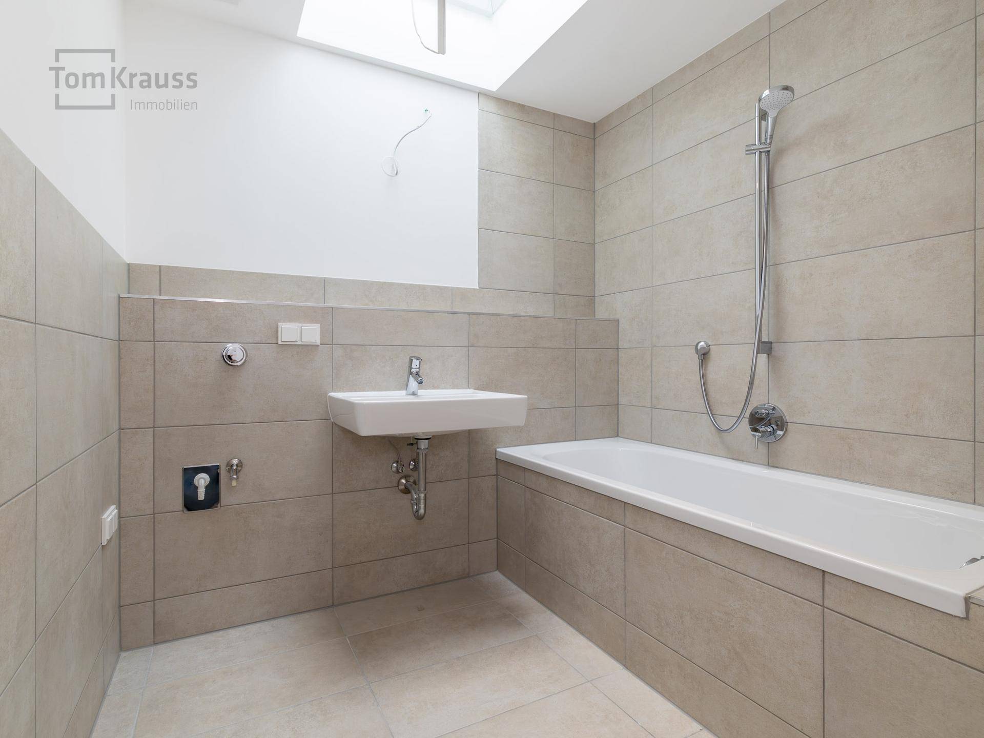 Modernes Badezimmer mit beigefarbenen Fliesen, Badewanne, wandhängendem Waschbecken und Dachfenster.