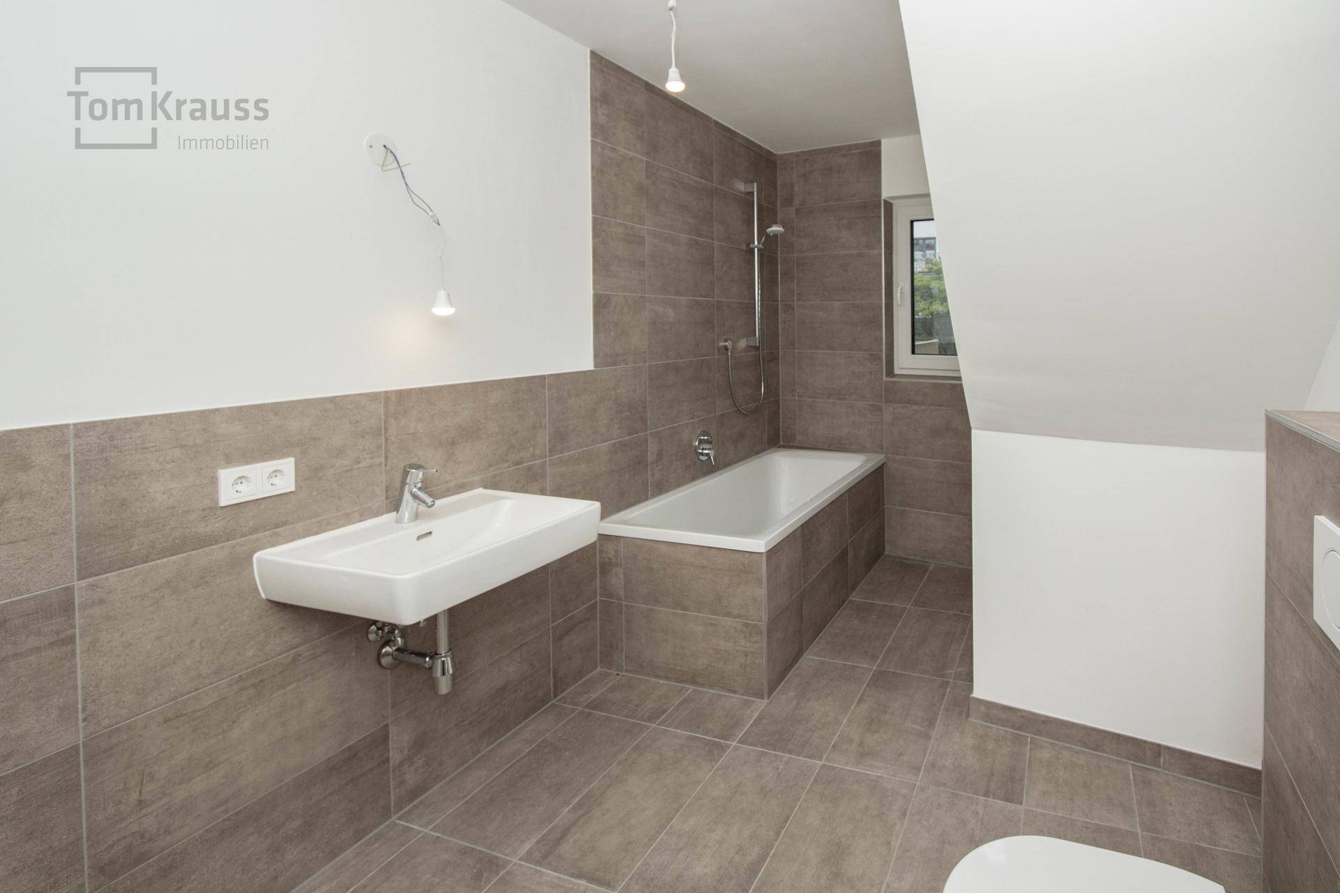 Modernes Badezimmer mit grauen Fliesen, Badewanne und Dachschräge, hell durch ein Fenster.