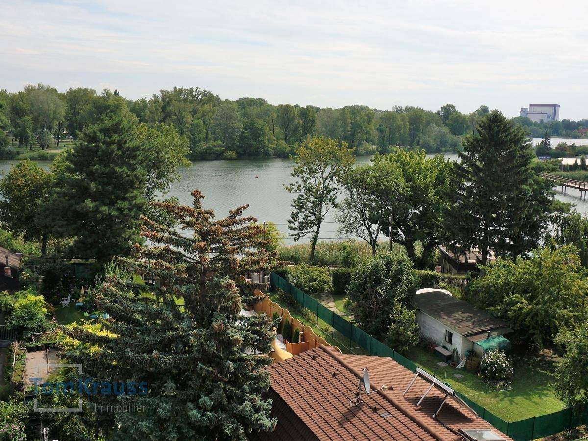 Panoramablick auf einen See mit üppiger Vegetation und einem Hausdach im Vordergrund.