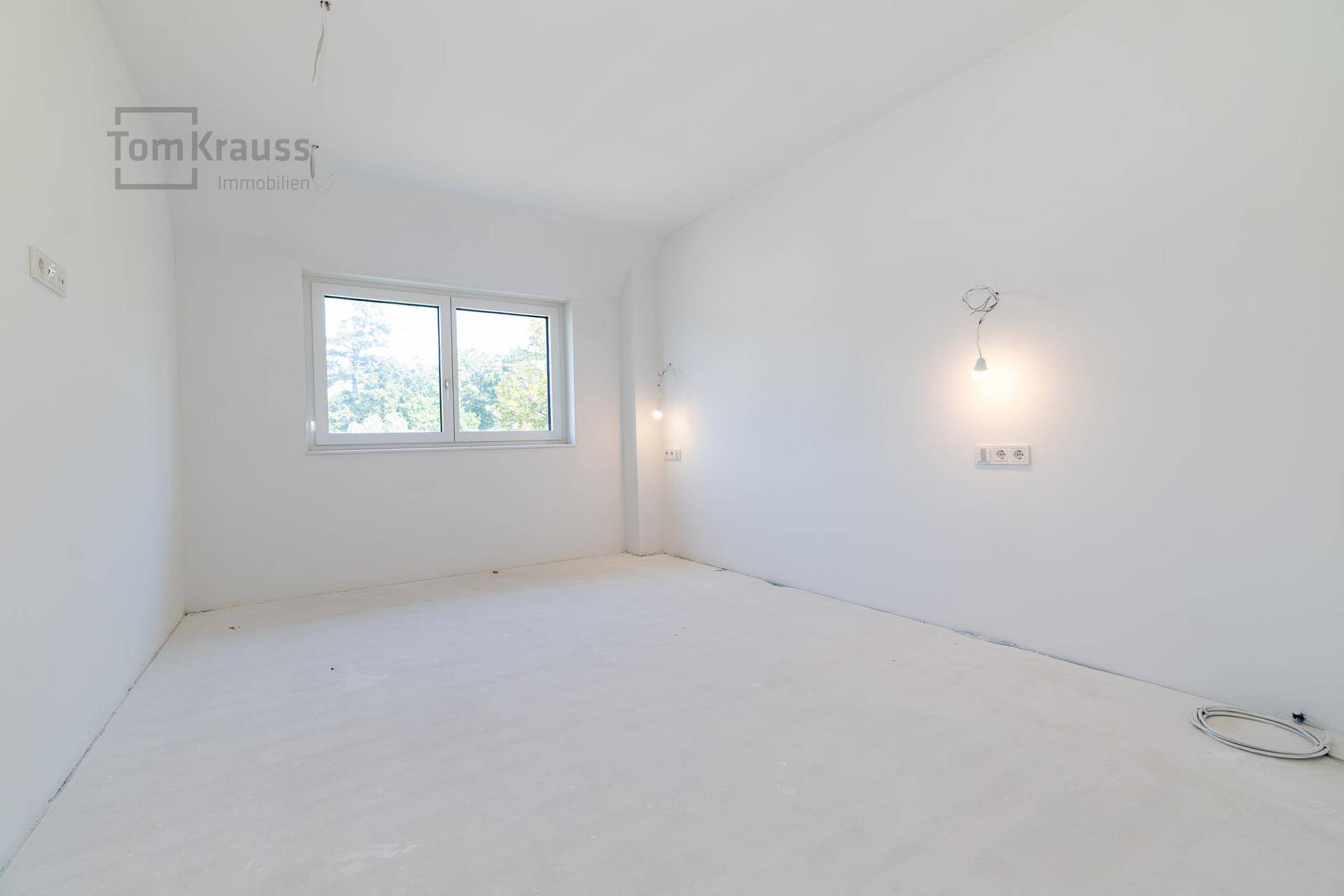 Leeres, helles Zimmer mit weiß gestrichenen Wänden und einem Fenster mit Blick ins Grüne.