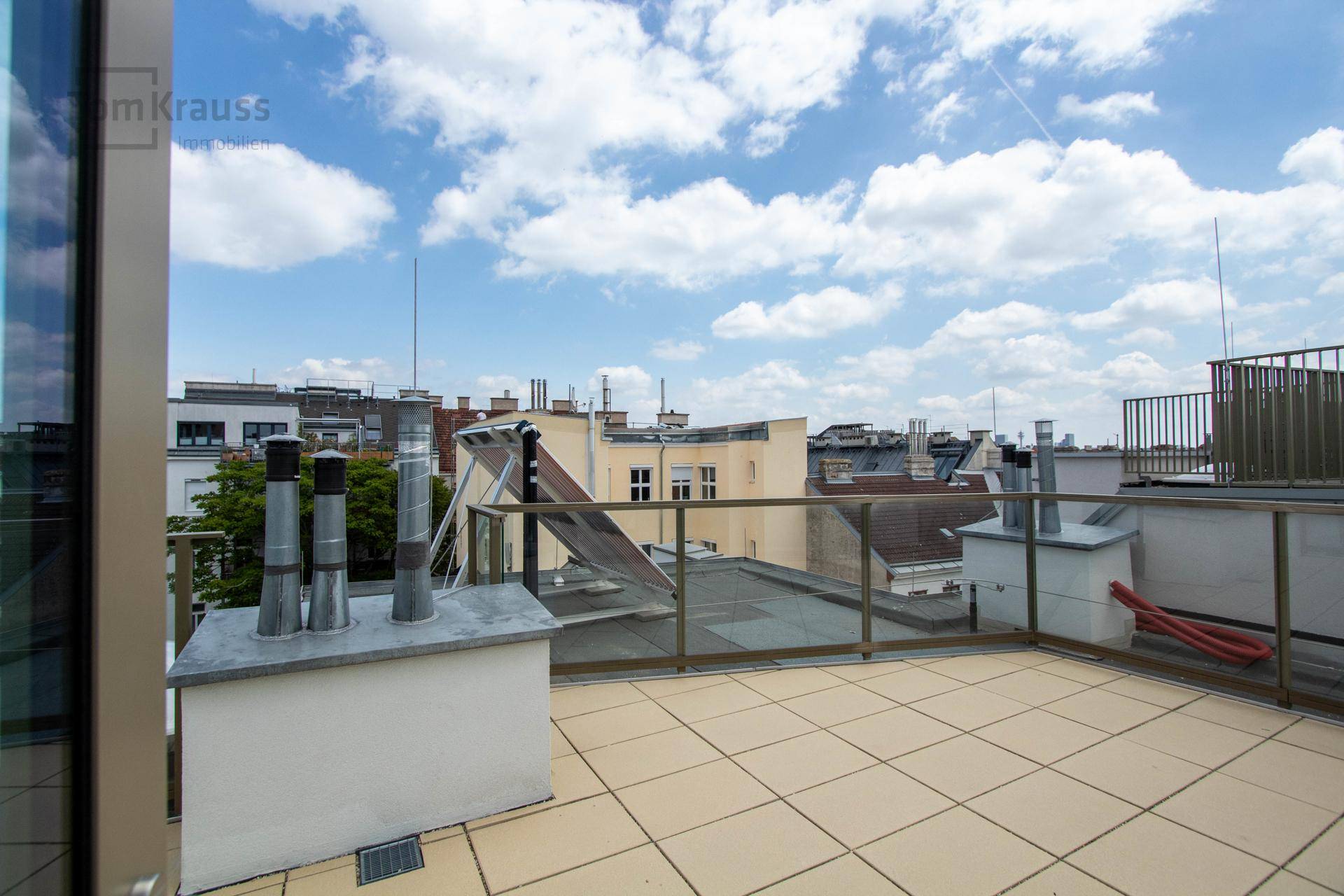 Dachterrasse mit hellen Fliesen, Glasgeländer und Blick auf die Dächer der Stadt.