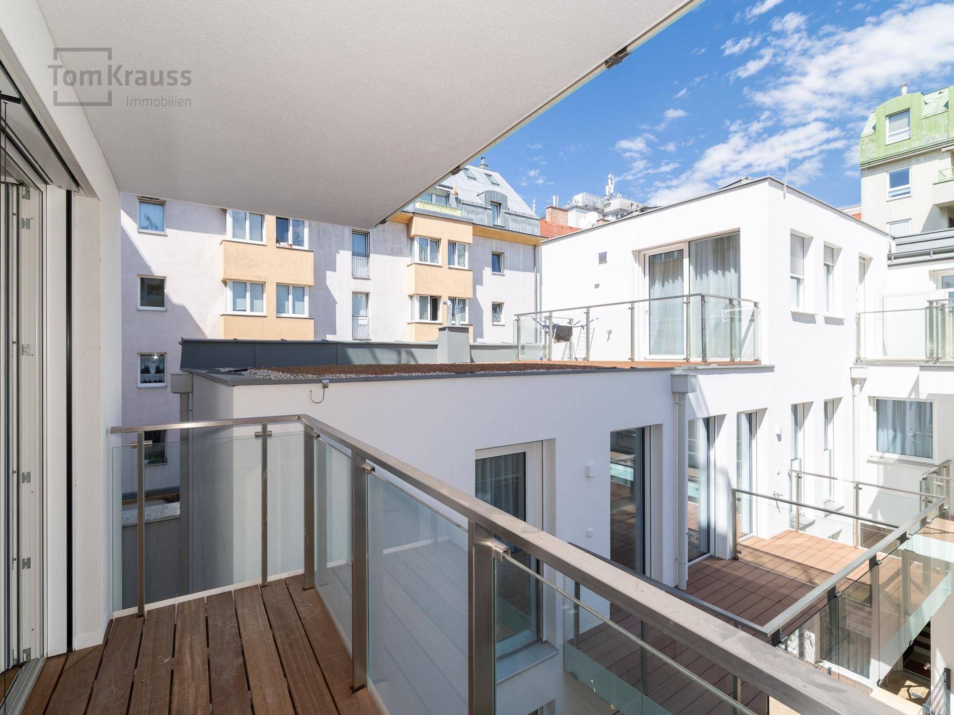 Balkon mit Holzbelag und Glasgeländer, bietet Ausblick auf den Innenhof und die umliegenden Gebäude.