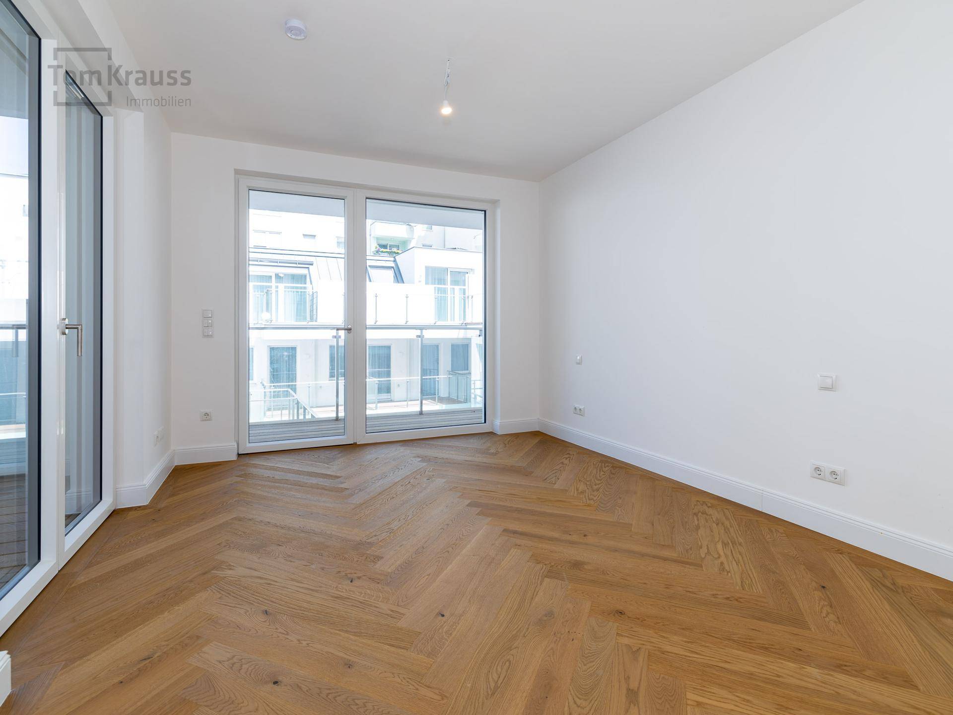 Lichtdurchflutetes Zimmer mit Fischgrätparkett und großen Fenstern, die auf einen Balkon führen.