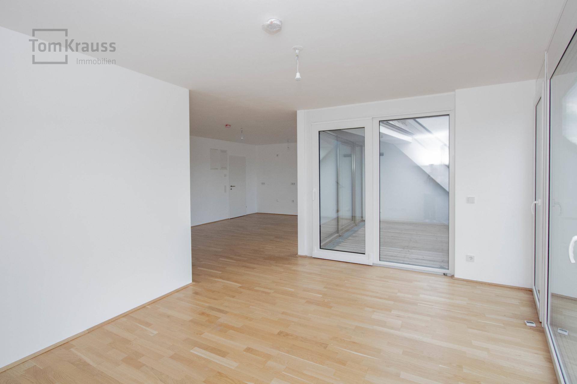 Heller, leerer Raum mit Holzboden und großen Glasfronten, die viel Licht hereinlassen.