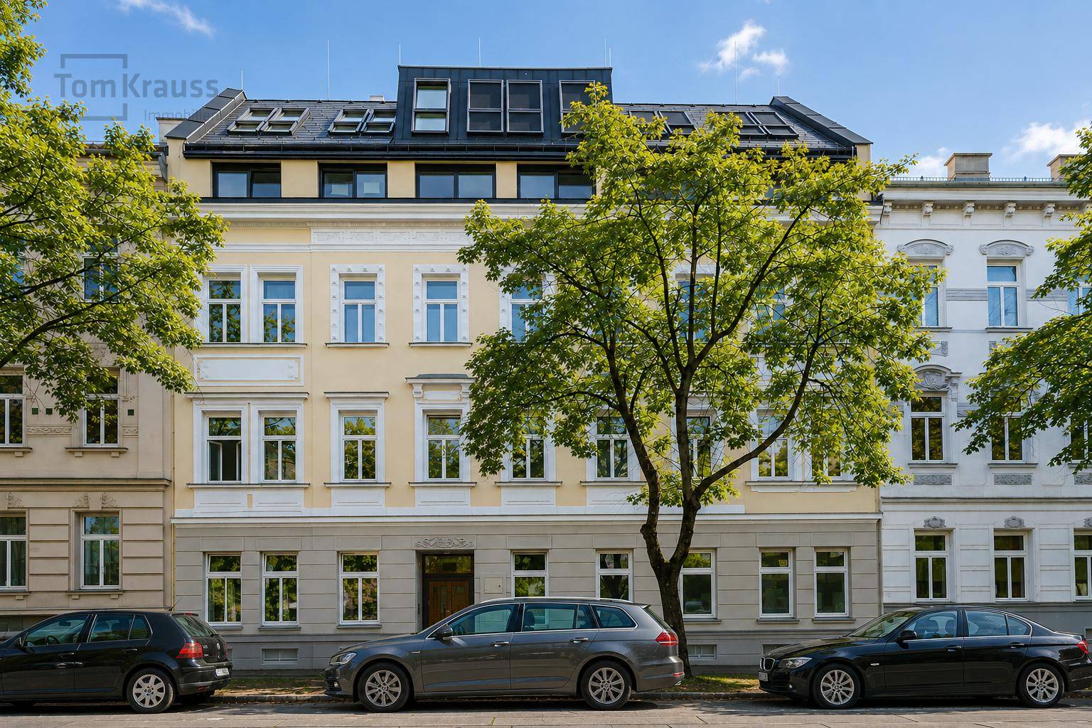 Klassische Fassade eines Mehrfamilienhauses mit vielen Fenstern und Bäumen davor an einer Straße.