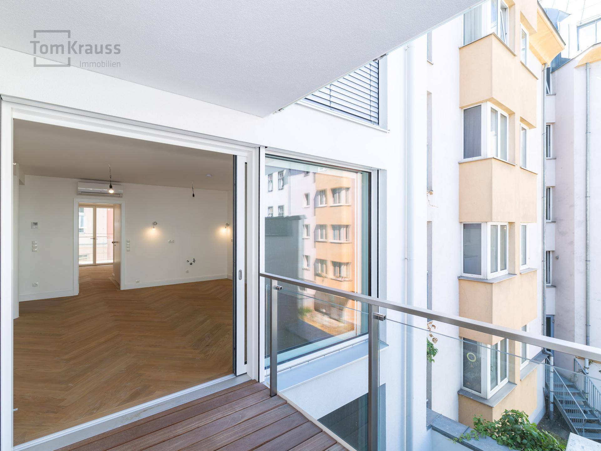 Balkon mit Holzbelag und Glasgeländer, bietet Blick auf den Innenhof und andere Balkone.