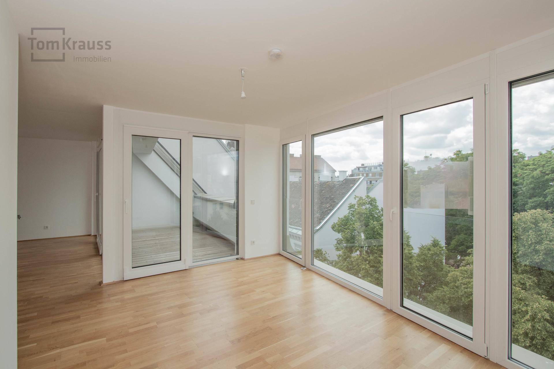 Heller, leerer Raum mit großen Fenstern, Holzboden und Zugang zu einem kleinen Balkon.