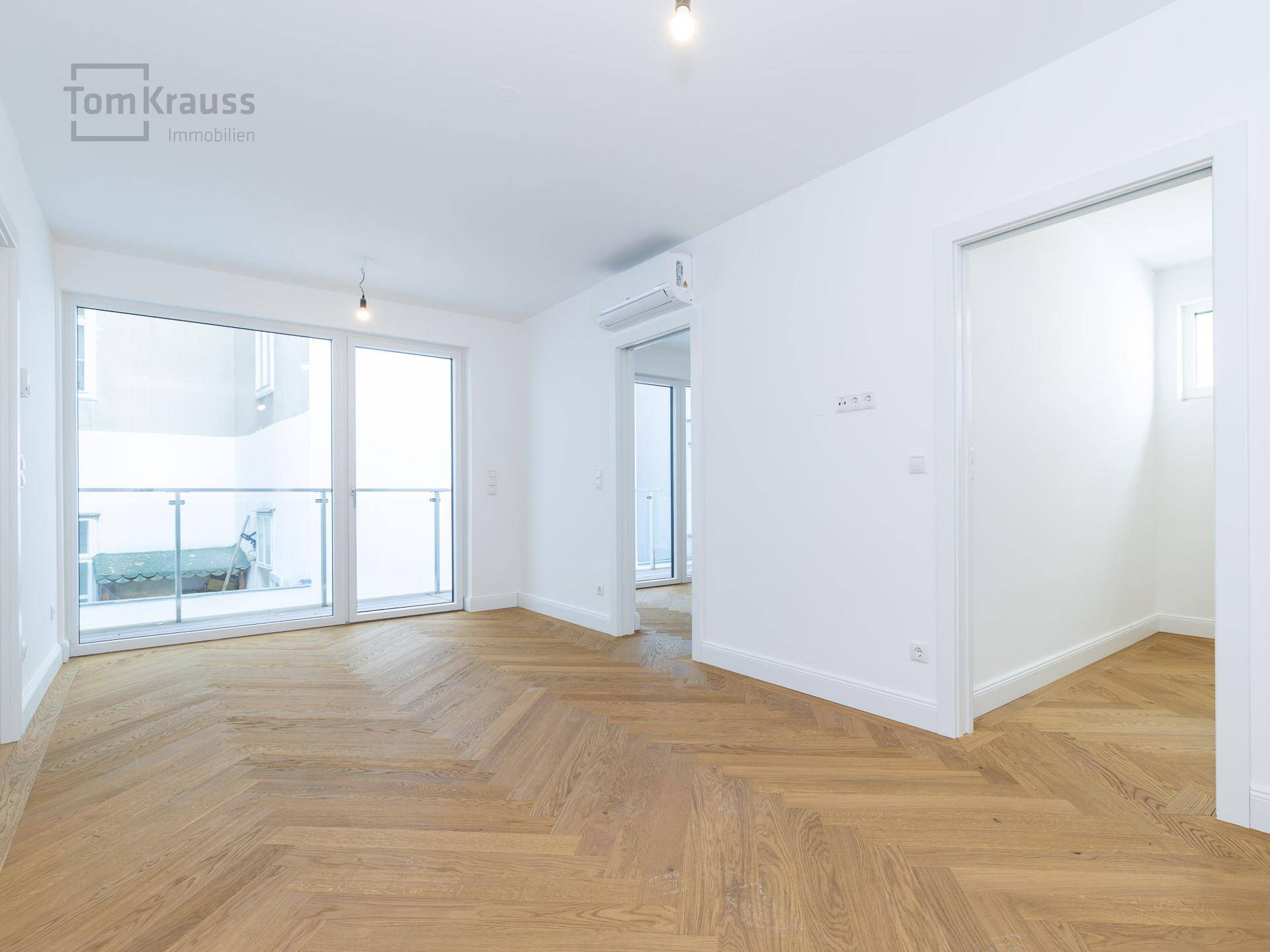 Geräumiges, leeres Wohnzimmer mit elegantem Fischgrätparkett und großen Fenstern zum Balkon.