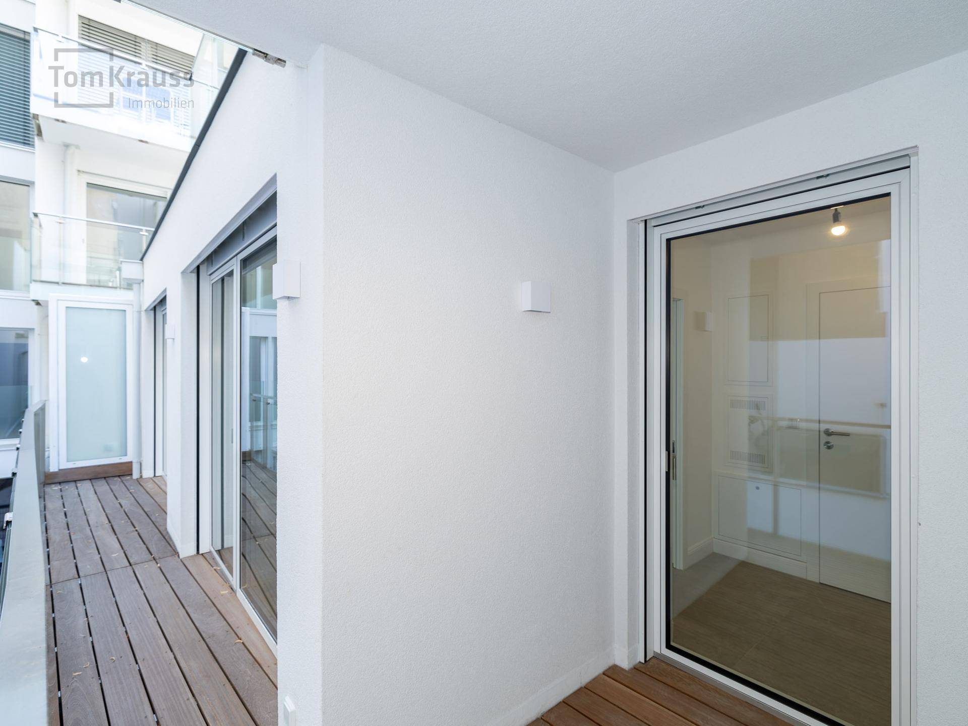 Schmaler Balkon mit Holzbelag und Glasgeländer, der mehrere Räume verbindet.