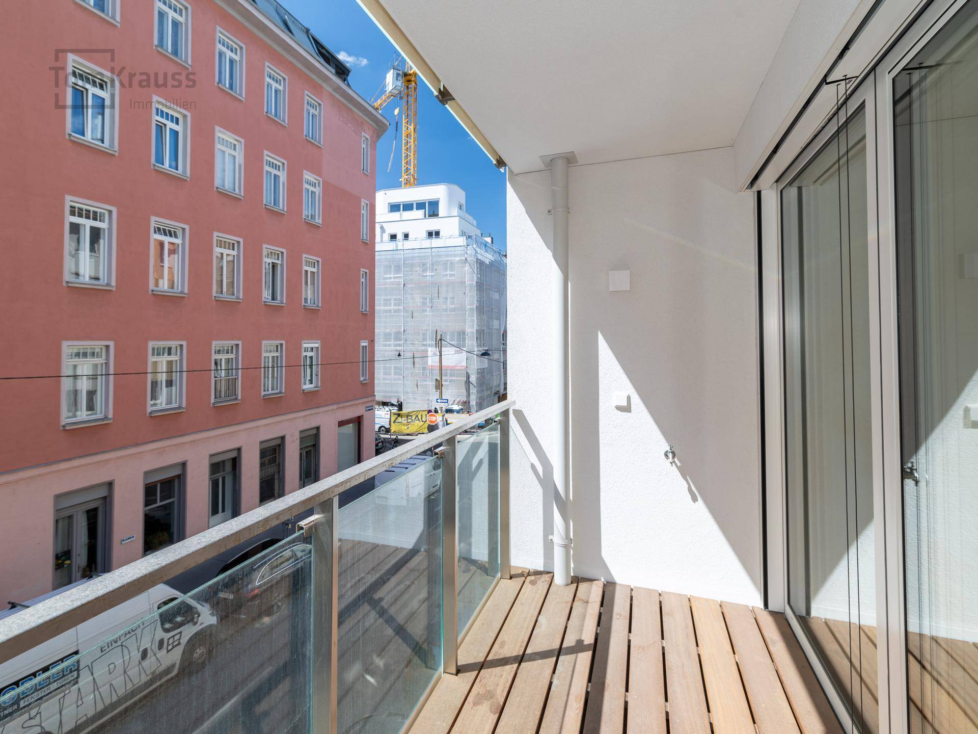 Kleiner Balkon mit Holzbelag und Glasgeländer, Blick auf die Straße und umliegende Gebäude.