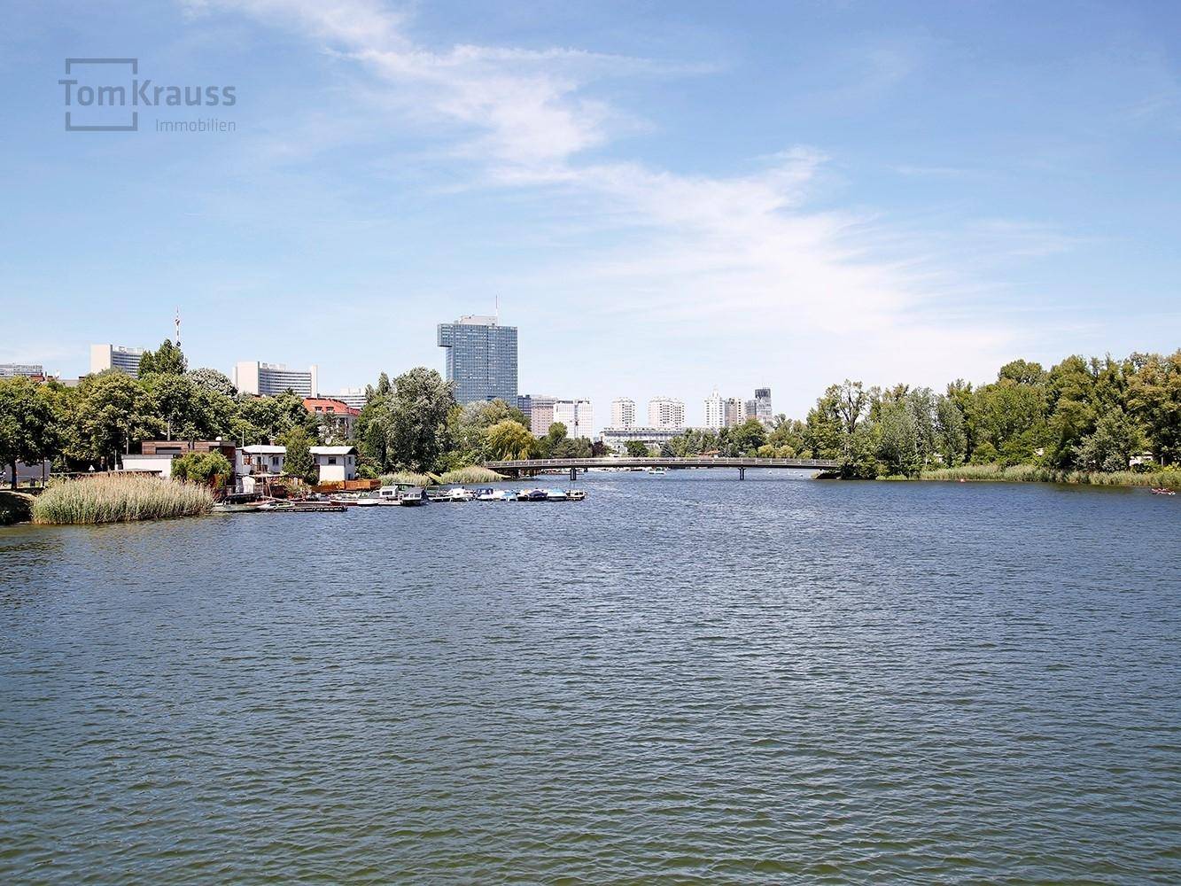 Weitläufiger Blick über einen See mit Stadtpanorama und grünen Ufern unter blauem Himmel.