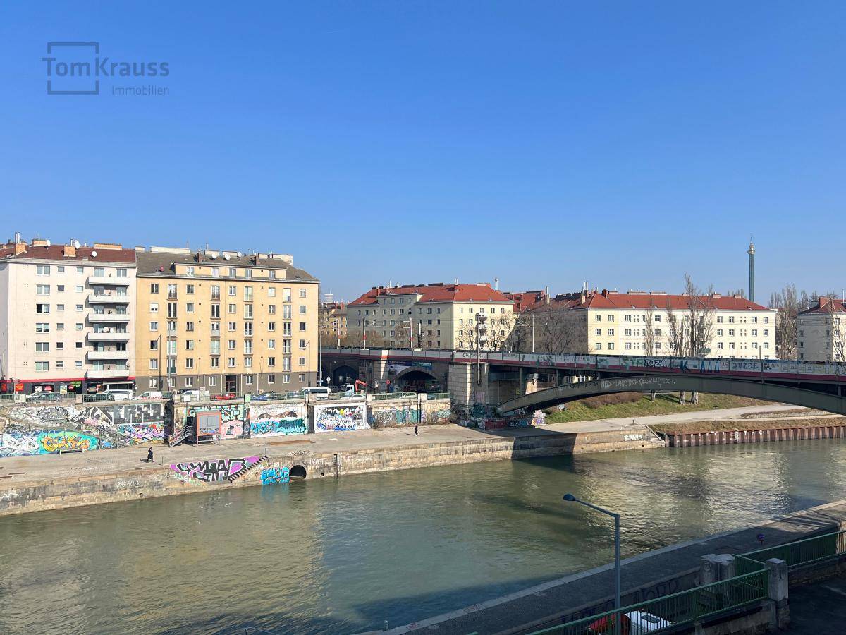 Weitläufiger Blick auf den Donaukanal mit Brücke und umliegenden Gebäuden bei Tageslicht.