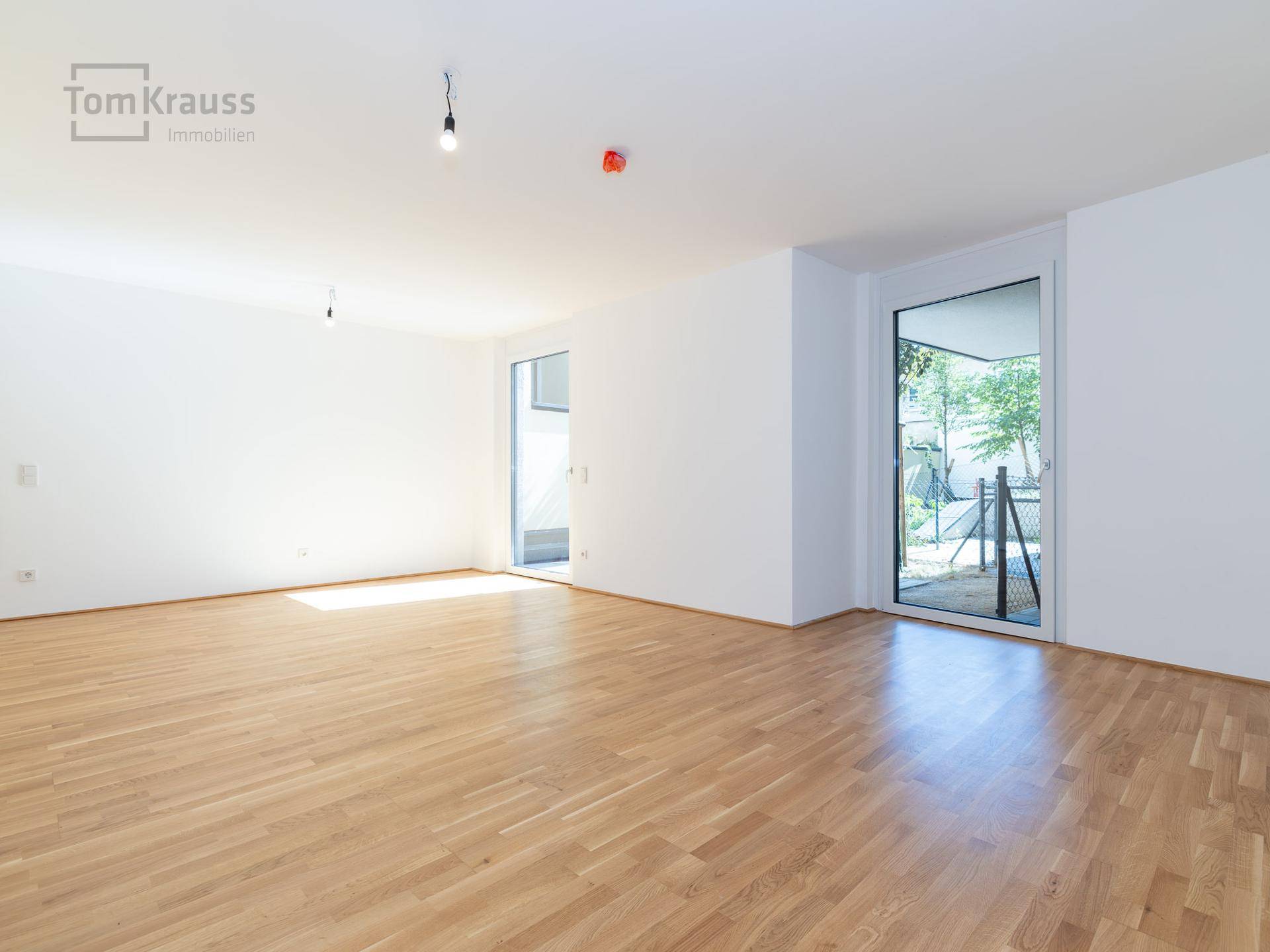Geräumiges, leeres Wohnzimmer mit hellem Holzboden und großer Glastür zum Garten oder zur Terrasse.