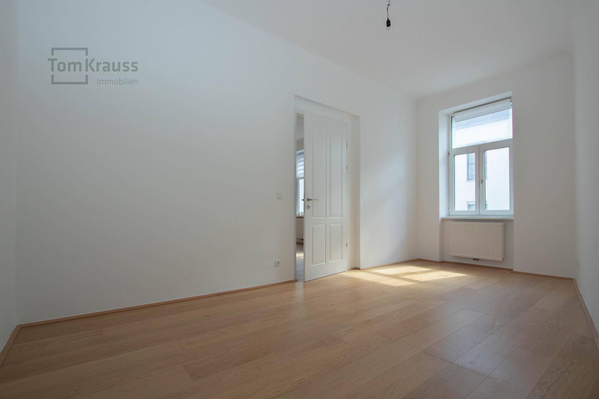 Leerer Raum mit hellem Parkettboden, weißen Wänden und einem Fenster mit Heizkörper.