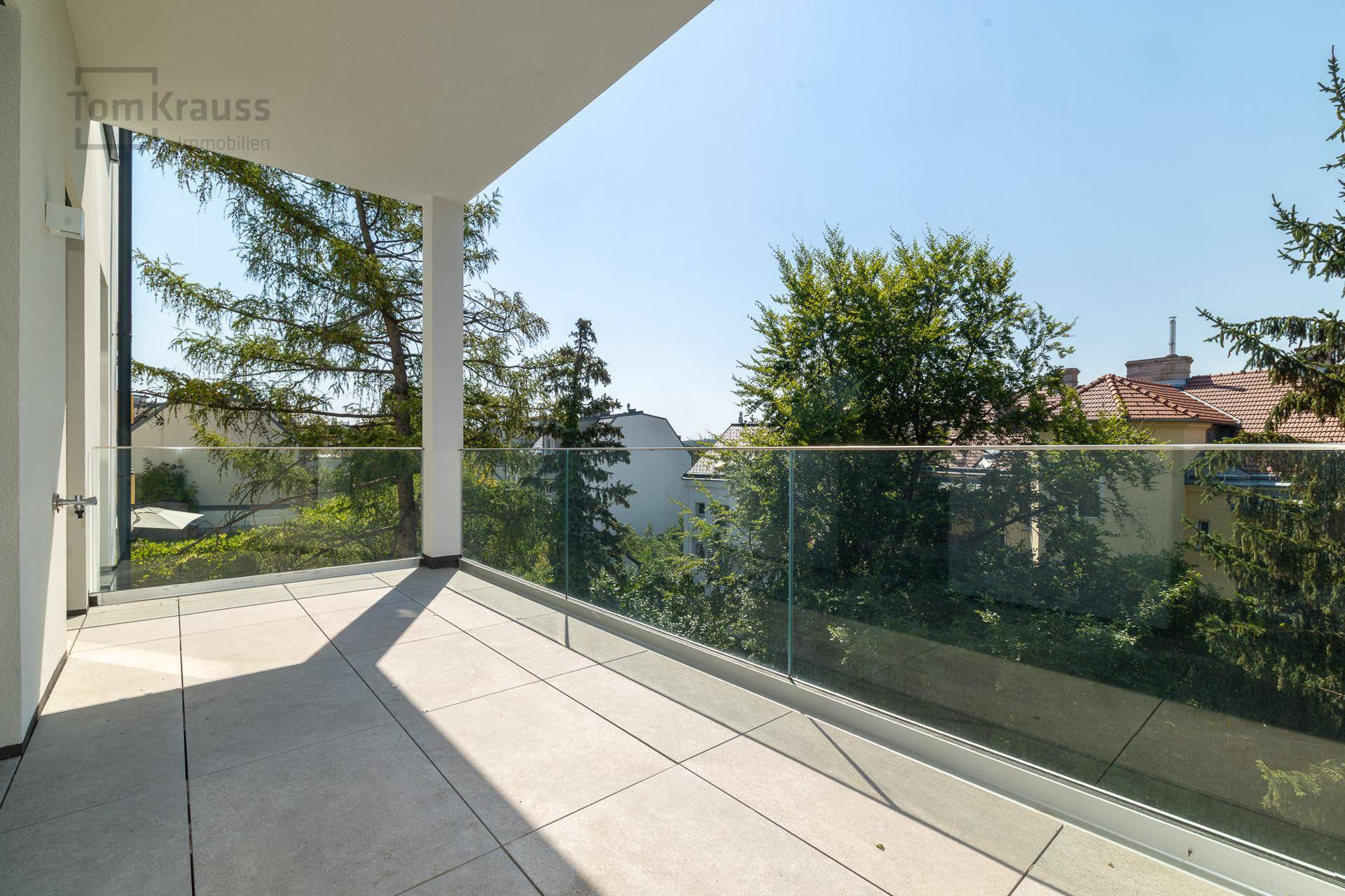 Geräumiger Balkon mit Fliesenboden und moderner Glasbrüstung, umgeben von Bäumen.