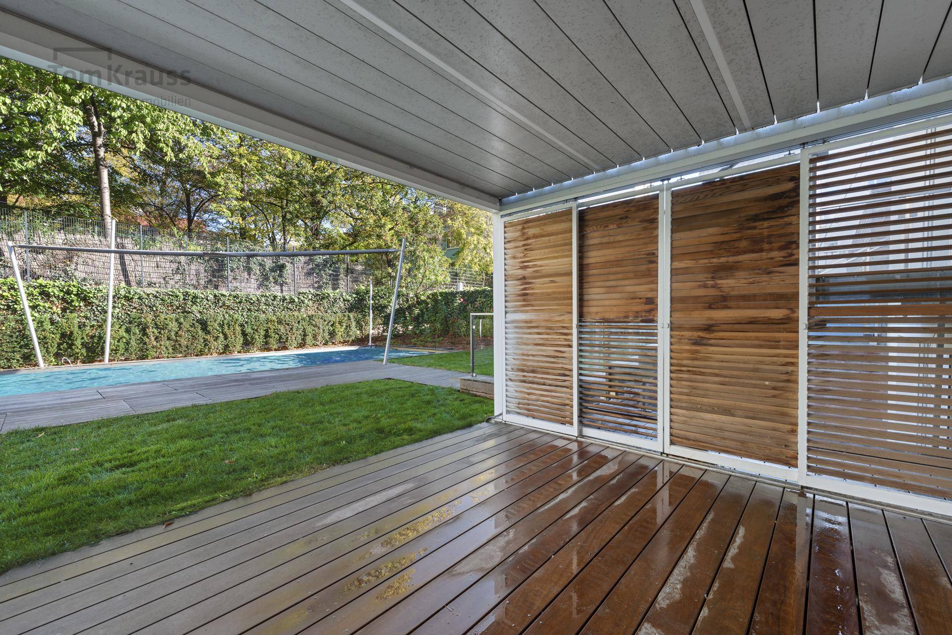Überdachter Außenbereich mit Holzterrasse, Rasenfläche und Volleyballfeld im Hintergrund.