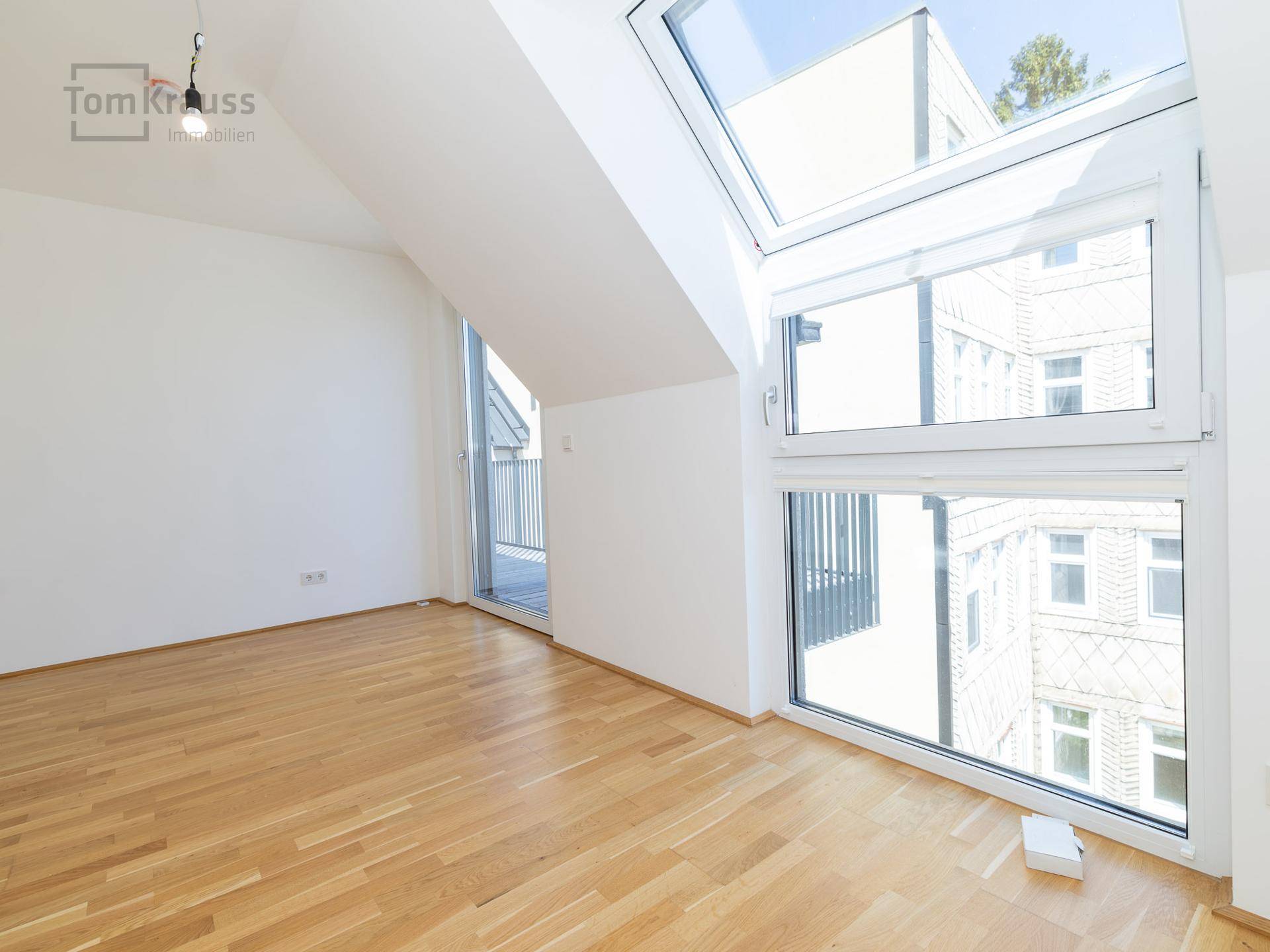 Helles, unmöbliertes Wohnzimmer mit Holzboden, großen Fenstern und Zugang zu einem Balkon.