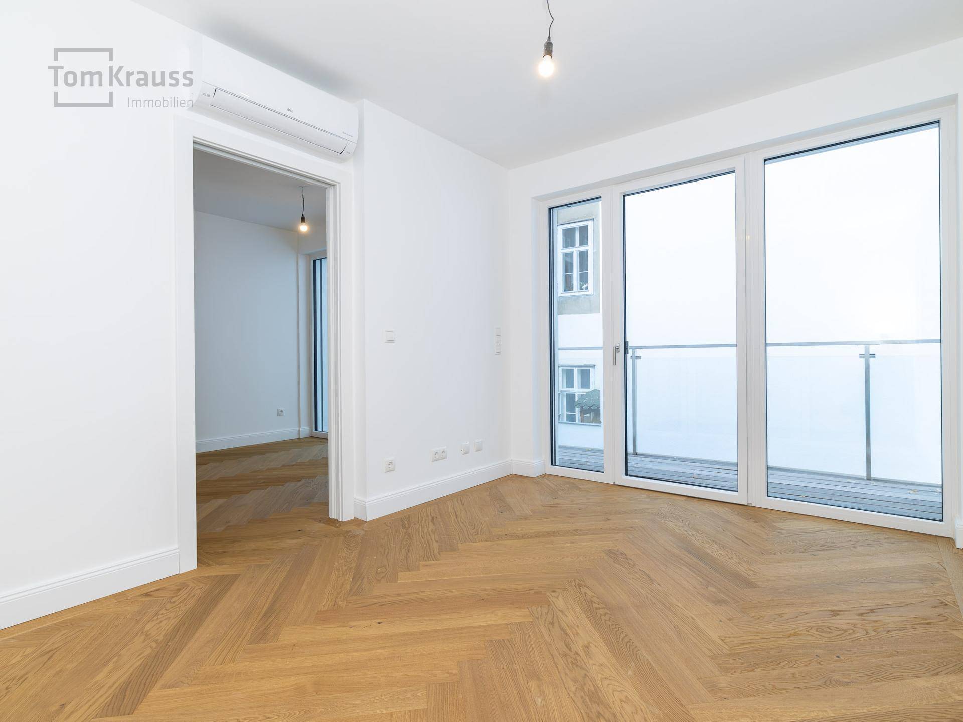 Leeres Schlafzimmer mit hellem Fischgrätparkett und großen Fenstern, die auf einen Balkon führen.