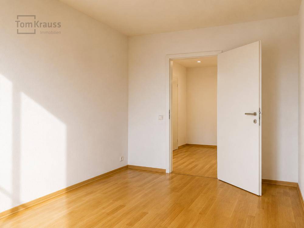 Leeres, helles Zimmer mit Holzboden und weiß gestrichenen Wänden, mit Blick in den Flur.