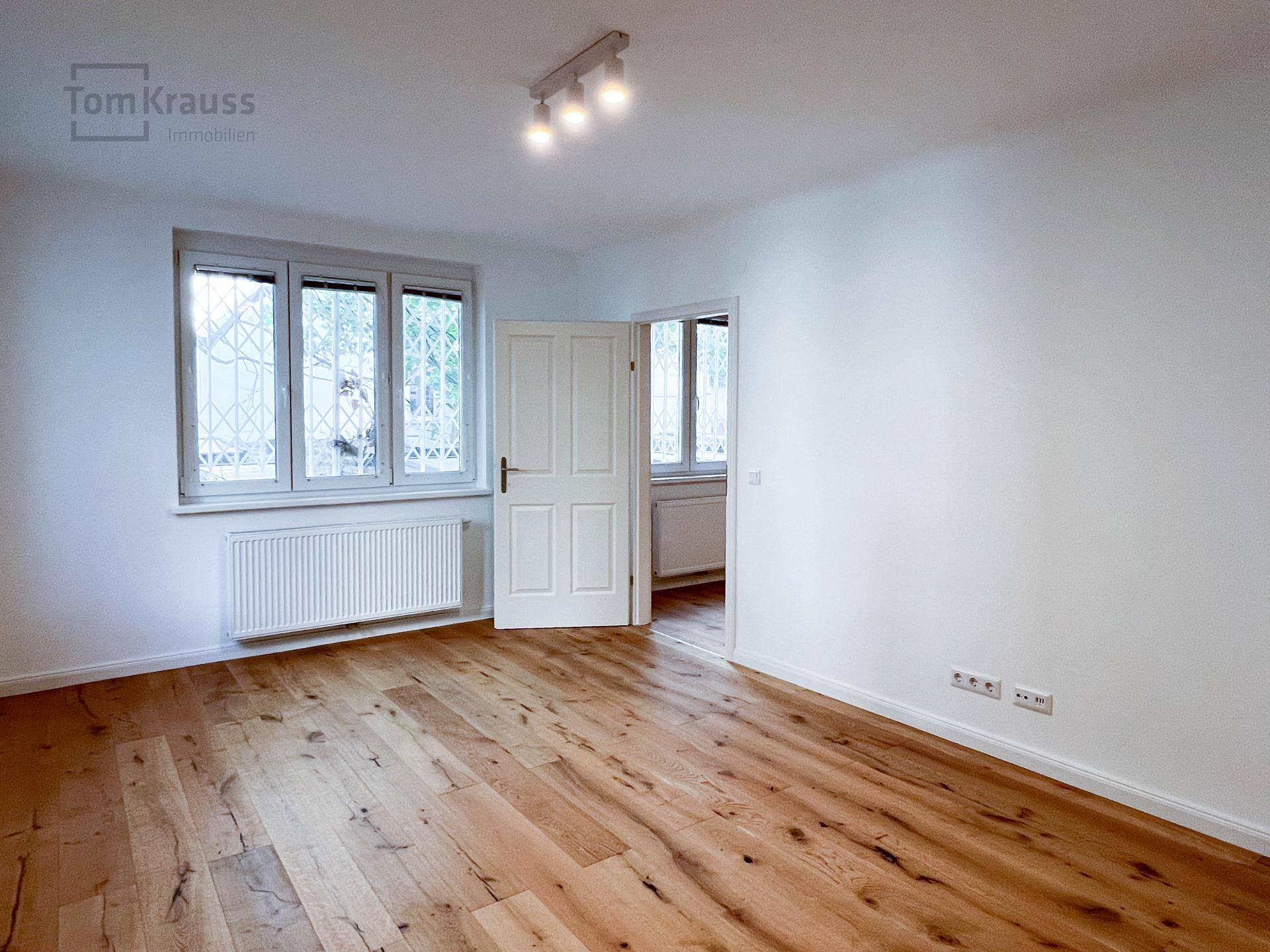 Freundliches Zimmer mit Holzboden, Fenster und Zugang zu einem weiteren Raum.