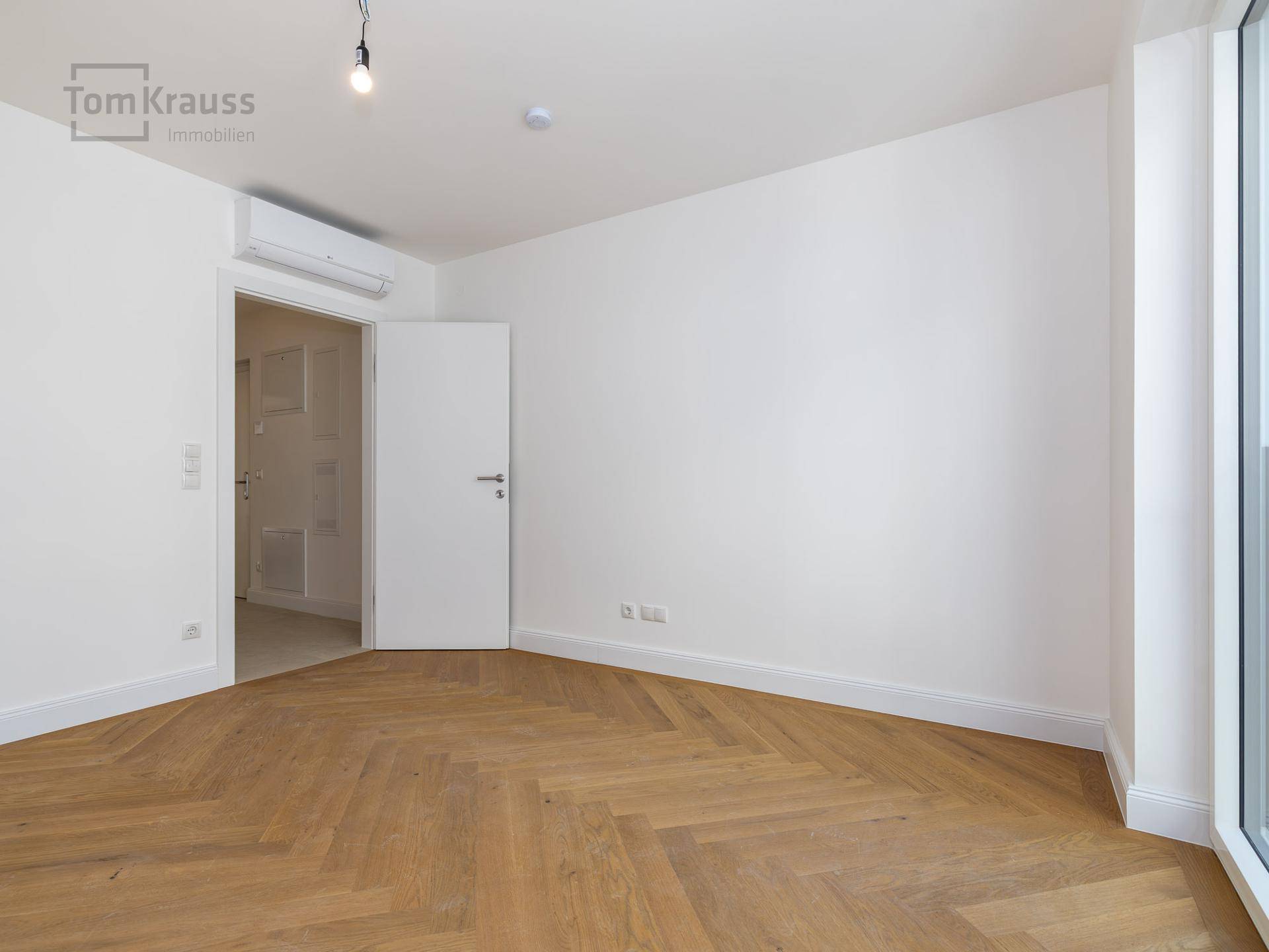 Leeres Zimmer mit Fischgrätparkett, weißen Wänden und Klimaanlage, bereit zum Einrichten.