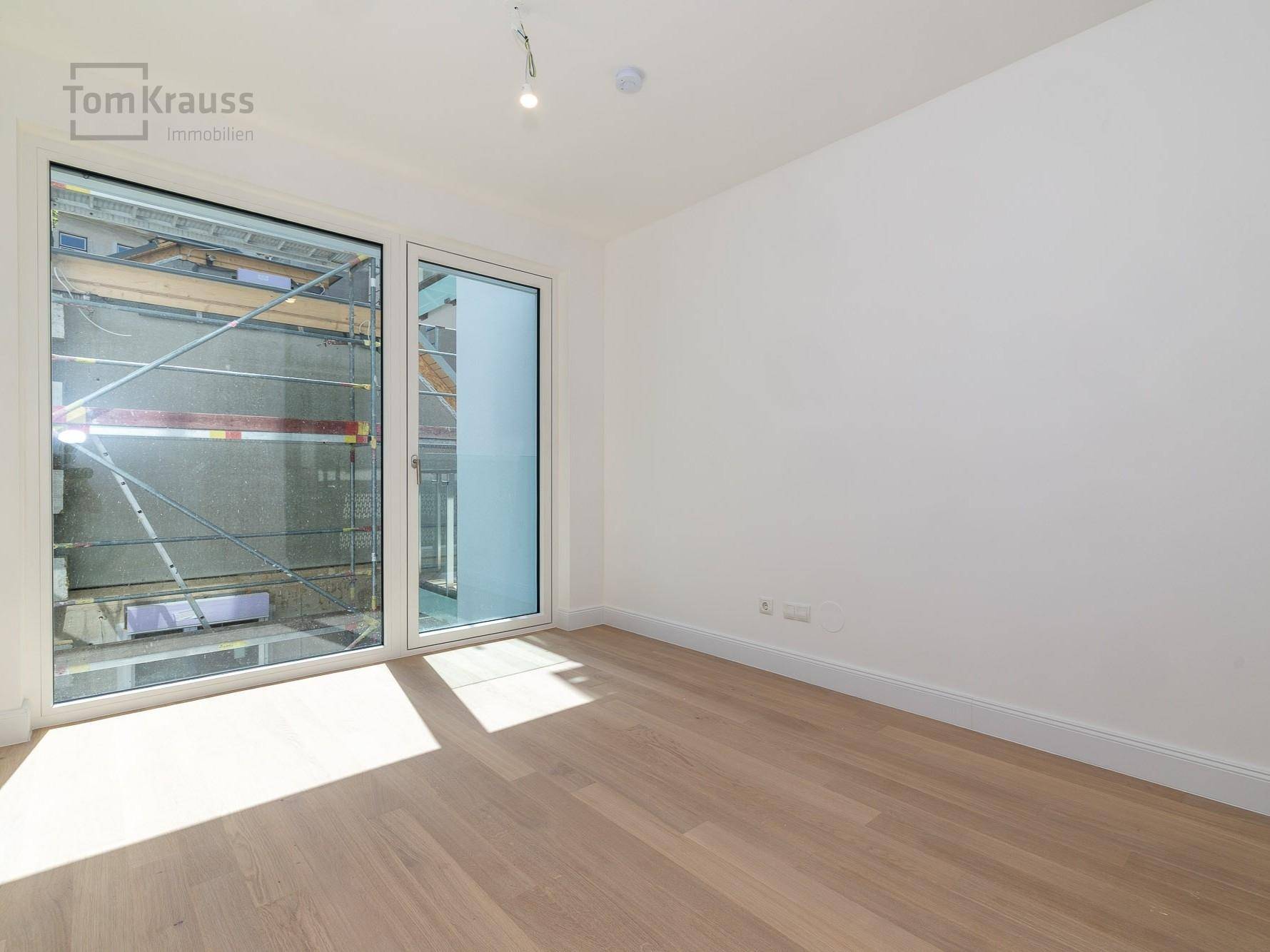 Leeres Zimmer mit hellem Holzboden und großen Fenstern, die viel Tageslicht hereinlassen.