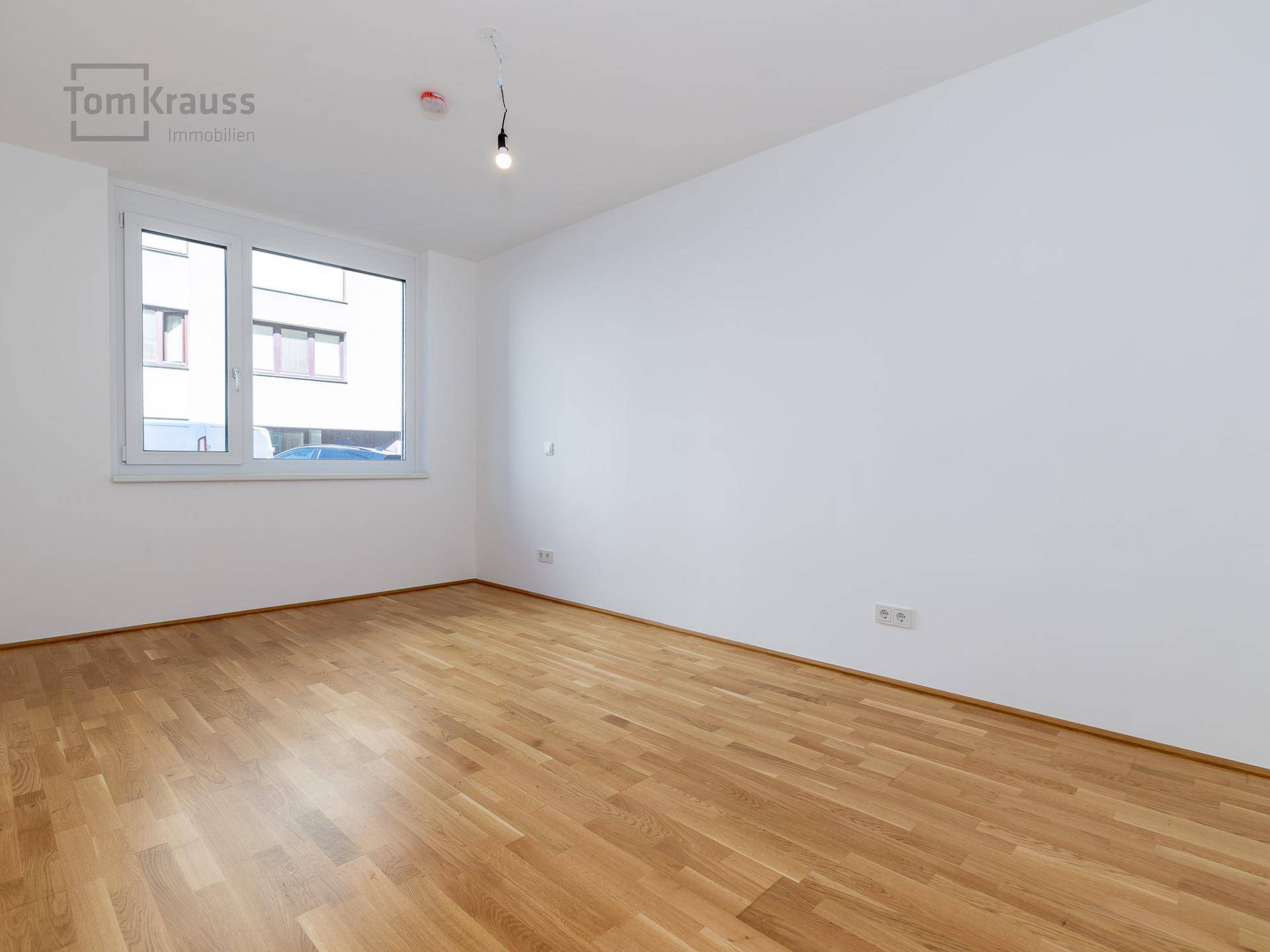 Leeres Zimmer mit hellem Holzboden und einem großen Fenster, das viel Tageslicht hereinlässt.