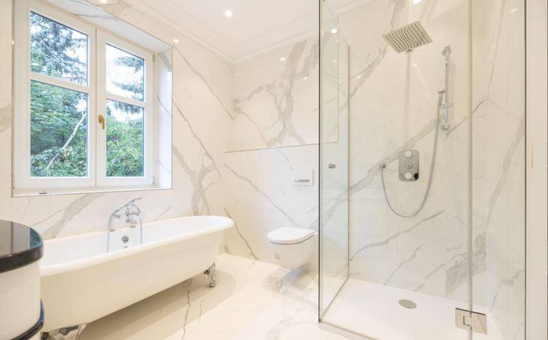 Luxuriöses Badezimmer mit freistehender Badewanne, begehbarer Dusche und Fenster mit Blick ins Grüne.