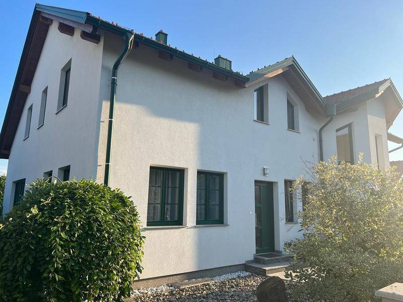 Seitenansicht des Hauses mit grünen Fensterrahmen, gepflegter Bepflanzung und sonnigem Ambiente.