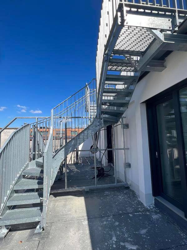 Eine moderne Metalltreppe führt auf eine Dachterrasse mit Zugang zu einem Gebäude.